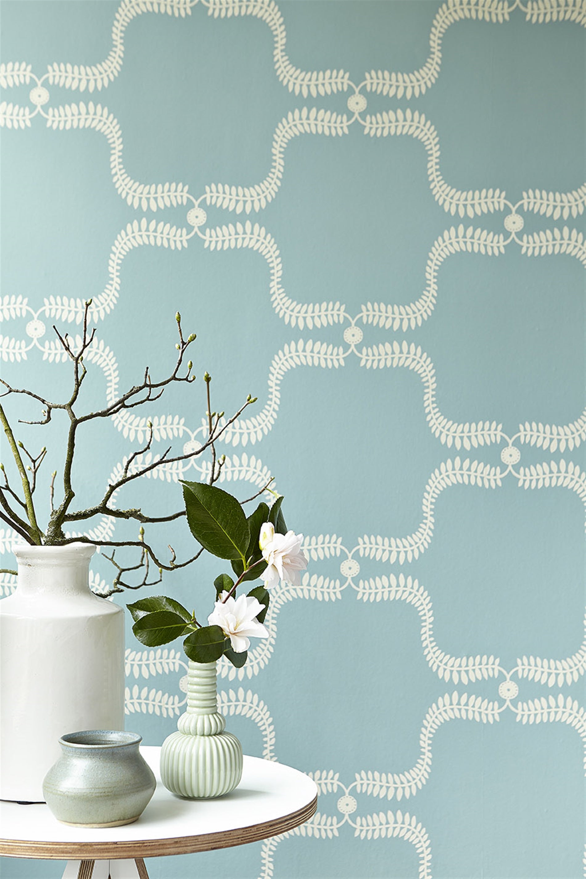 An end table with flowers stands in front of a wall papered in a leafy lattice print in cream on a light blue field.