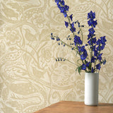 A vase of flowers stands in front of a wall papered in a playful wren and flower print in tan and cream.