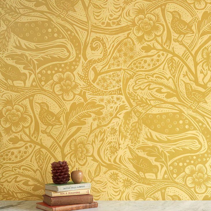 A stack of books stands in front of a wall papered in a playful wren and flower print in yellow and gold.