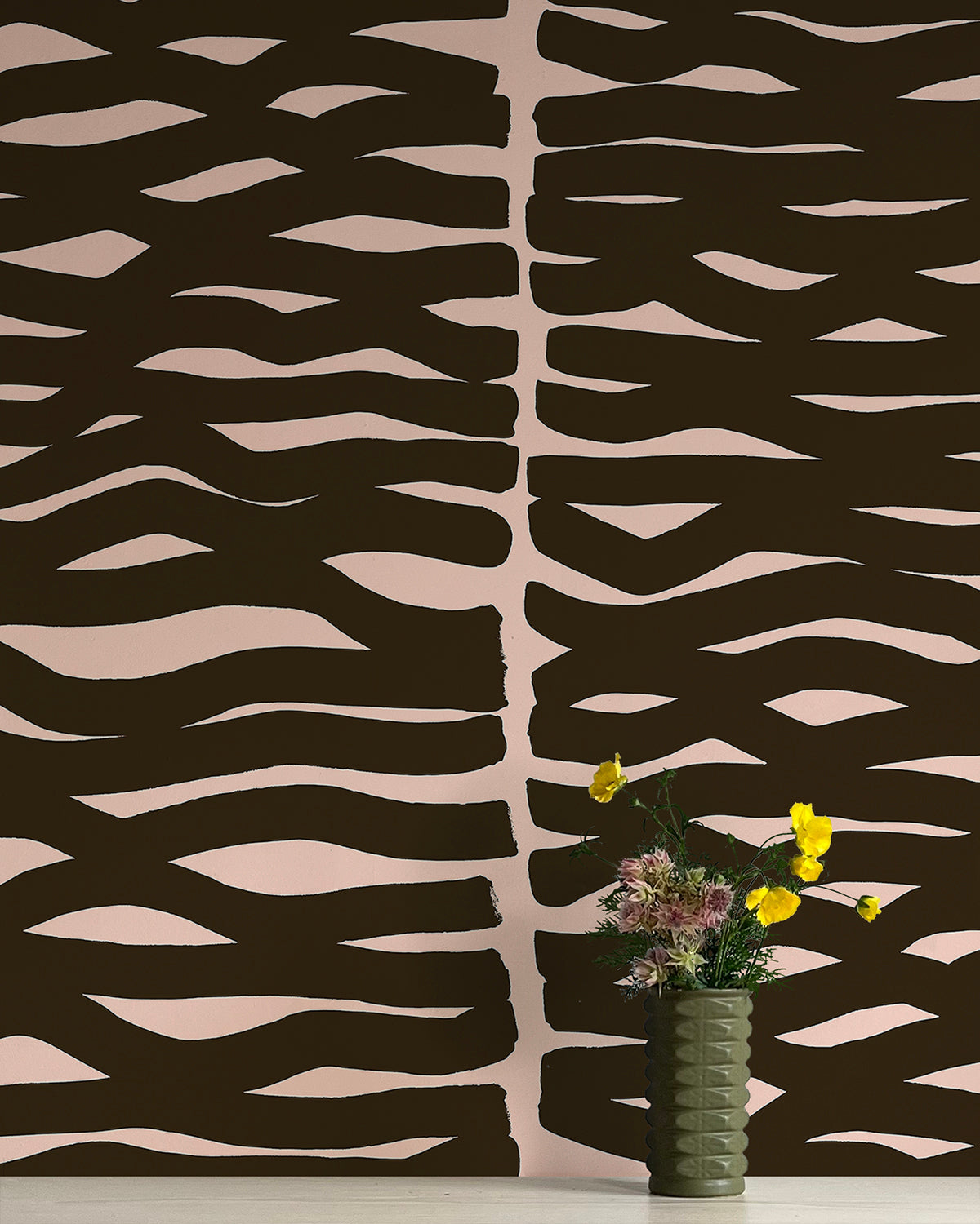 A vase of flowers stands in front of a wall papered in an abstract zigzag pattern in black on a light pink field.