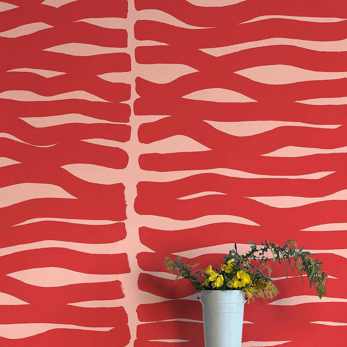 A vase of flowers stands in front of a wall papered in an abstract zigzag pattern in red on a light pink field.