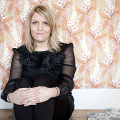 A blonde woman sitting on the ground in front of a densely patterned floral wallpaper, wearing all black with her hands around her knees.