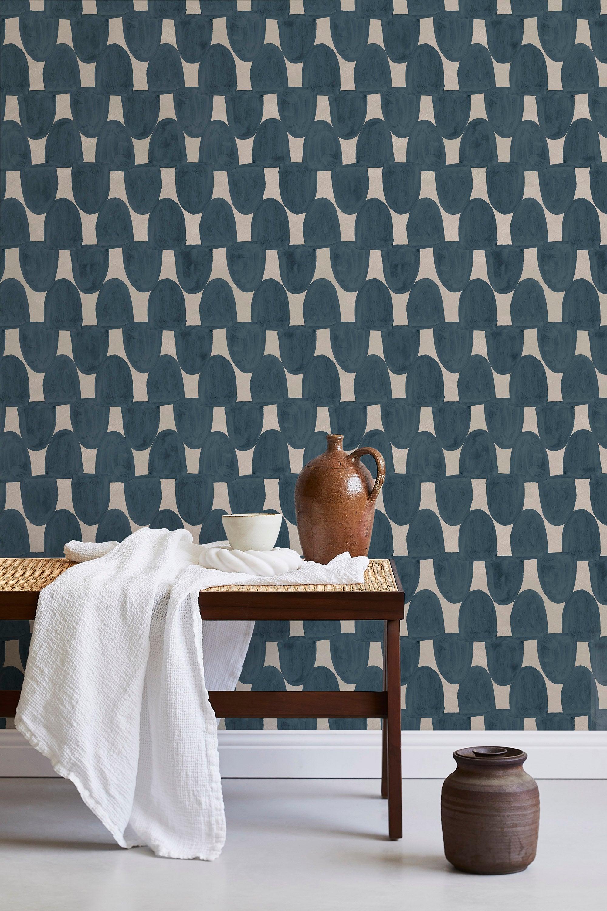 A bench with a towel and clay vases stands in front of a wall papered in an abstract scalloped print in navy and greige.