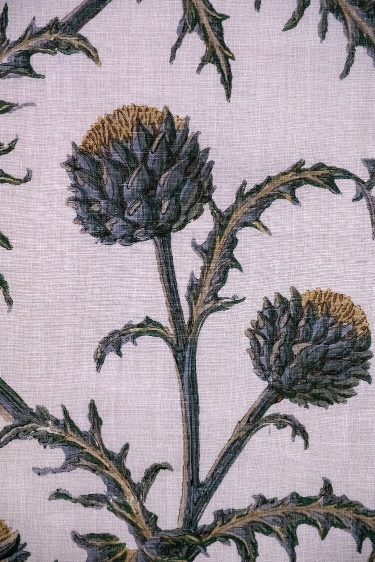 Detail of fabric in a repeating artichoke print in shades of purple, brown and green on a light purple field.