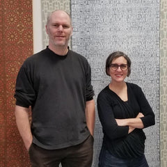 A man and woman dressed in black and dark gray, smiling and standing in front of a wall papered in several different geometric prints.