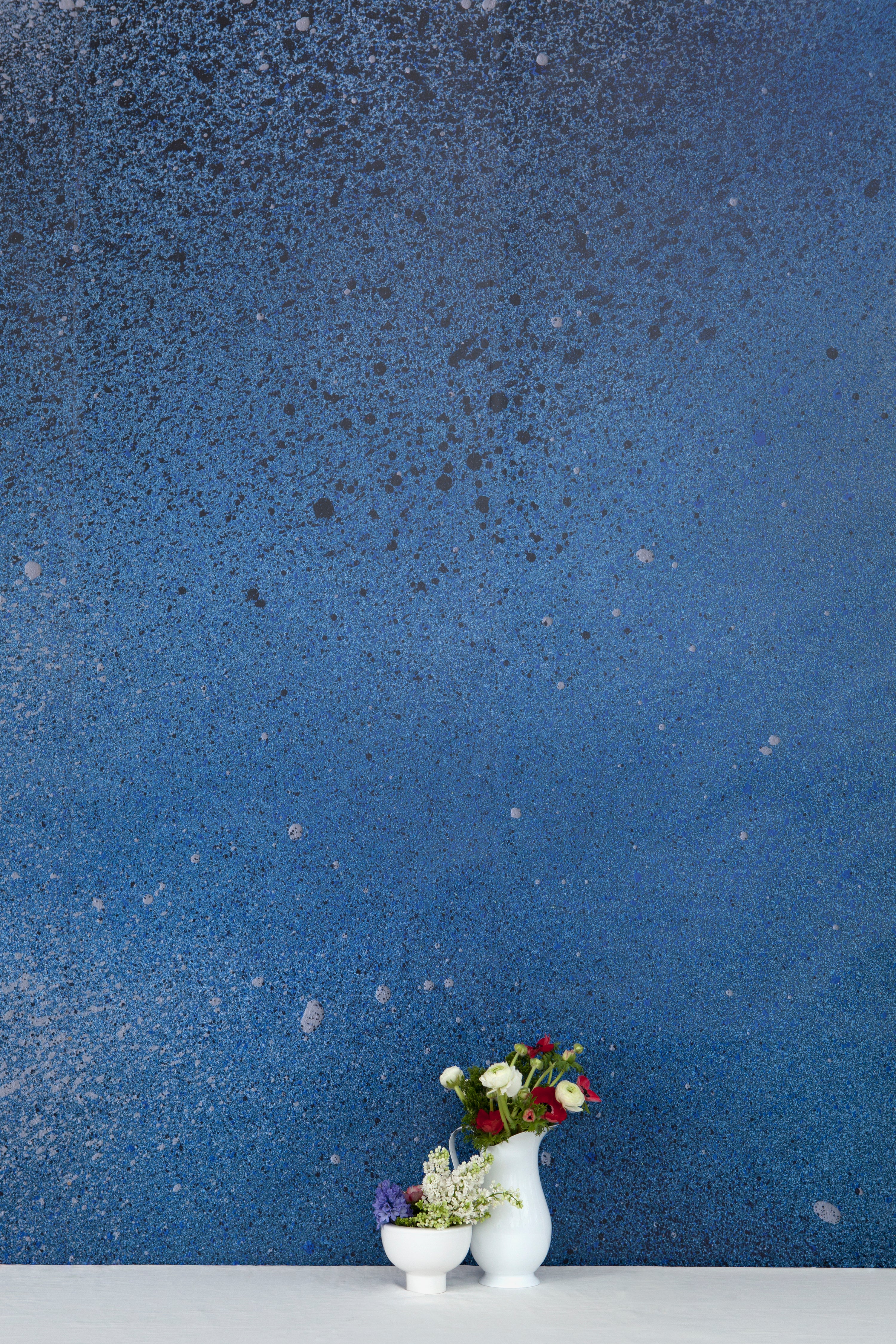 Two vases of flowers stand in front of a wall covered in an abstract ombré paint splatter print in blue, white and navy.