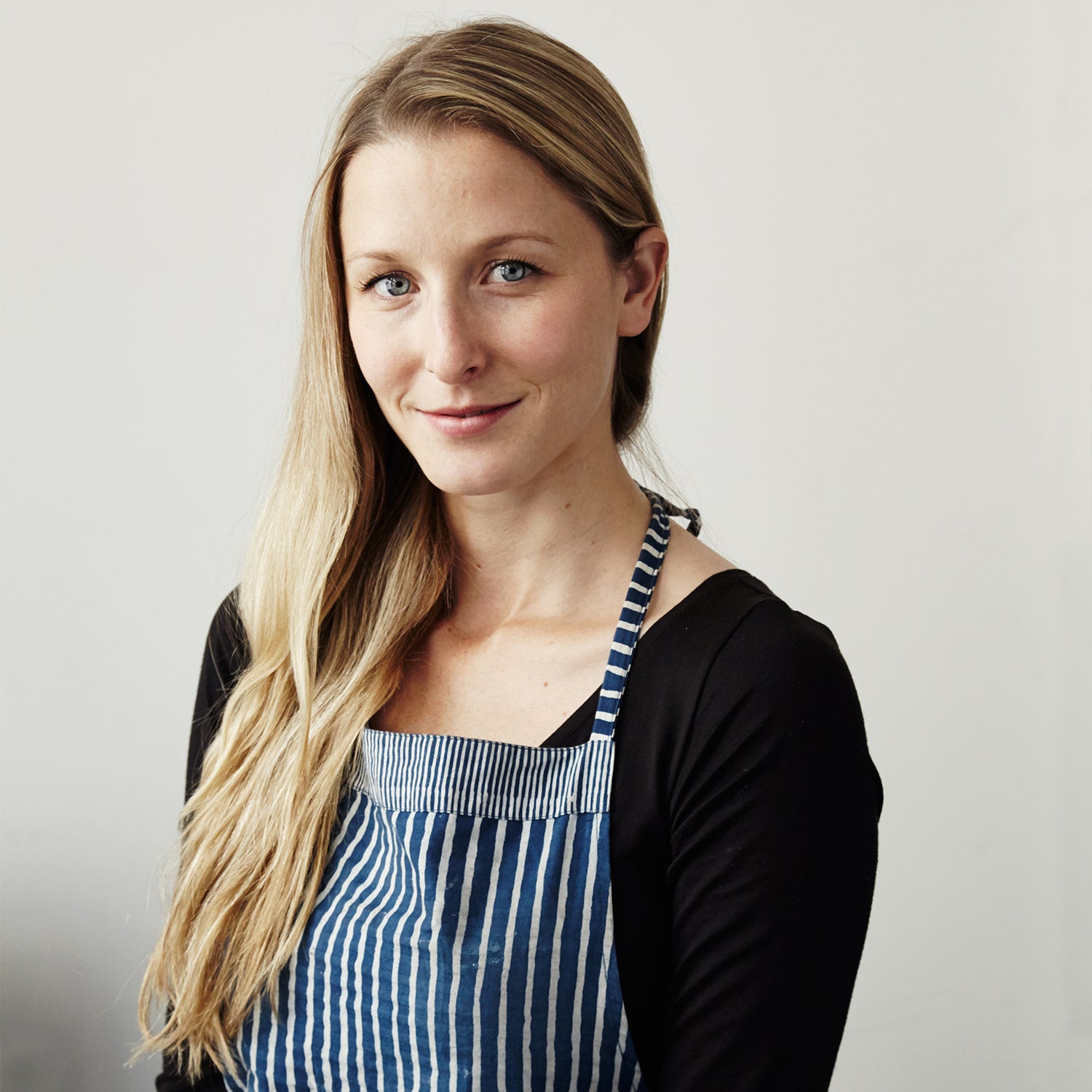 Close-up of a woman with long blonde hair smiling at the camera wearing a black top and a striped navy and white apron.