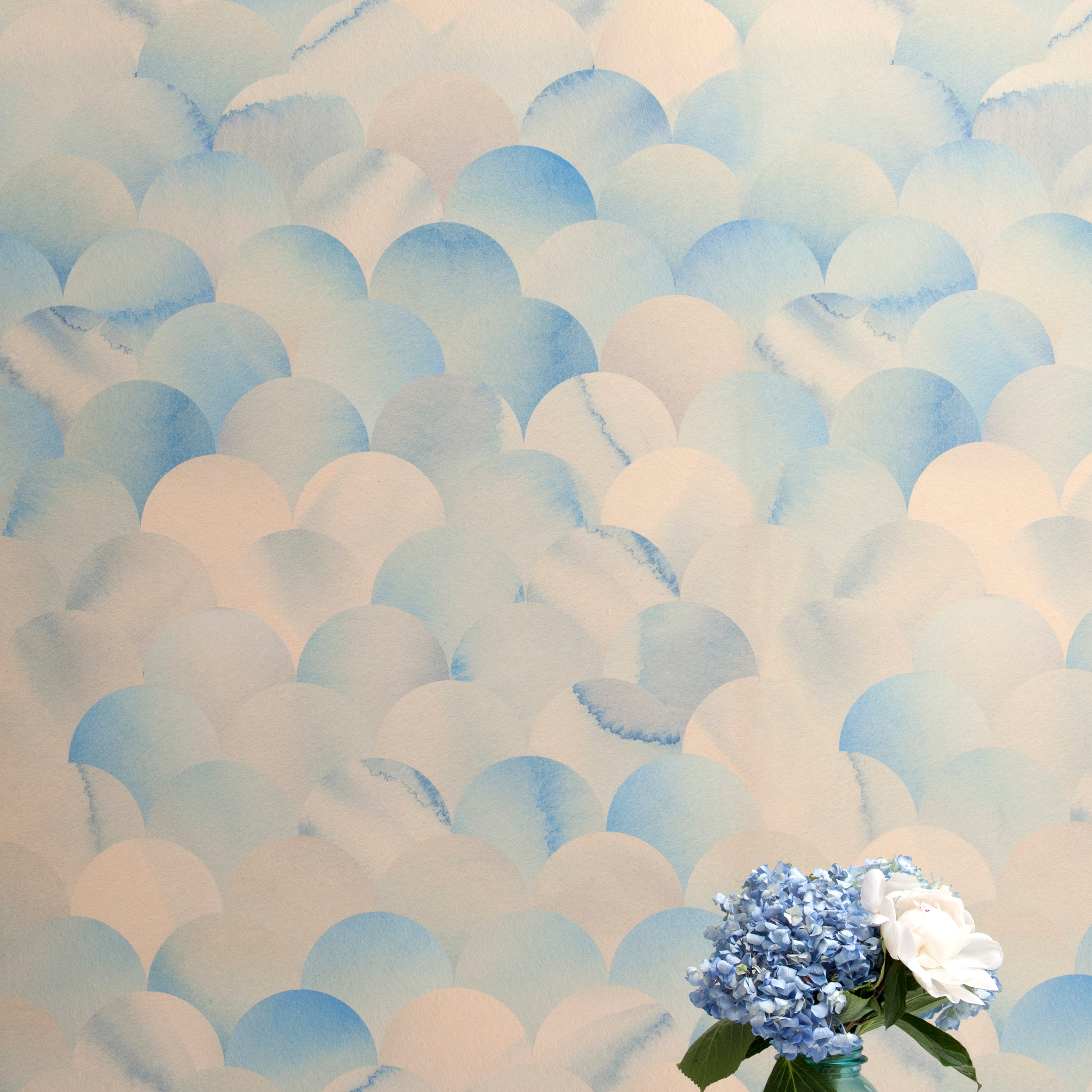 A vase of flowers stands in front of a wall papered in a repeating scalloped print in shades of blue and tan.