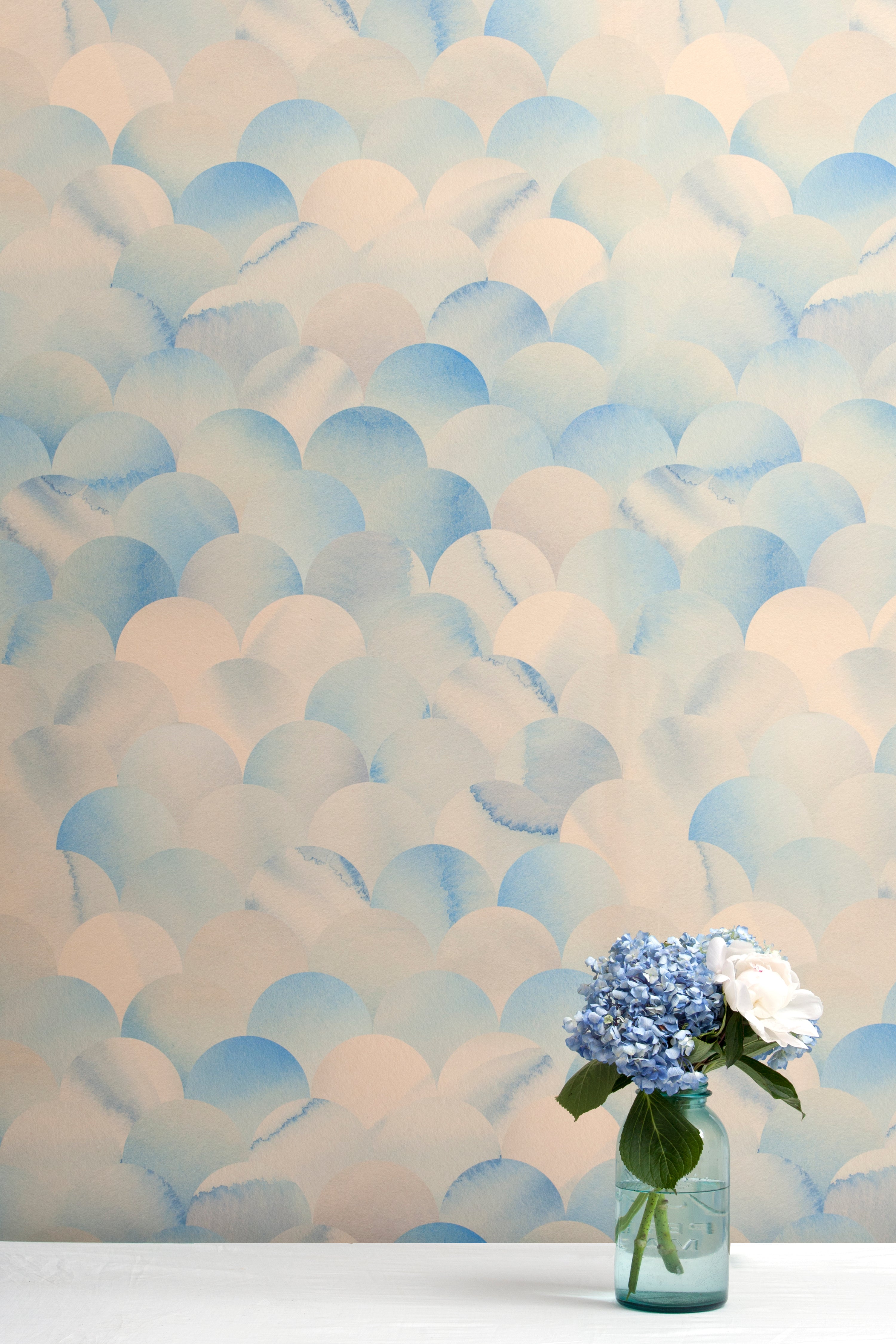 A vase of flowers stands in front of a wall papered in a repeating scalloped print in shades of blue and tan.