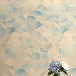 A vase of flowers stands in front of a wall papered in a repeating scalloped print in shades of blue and tan.