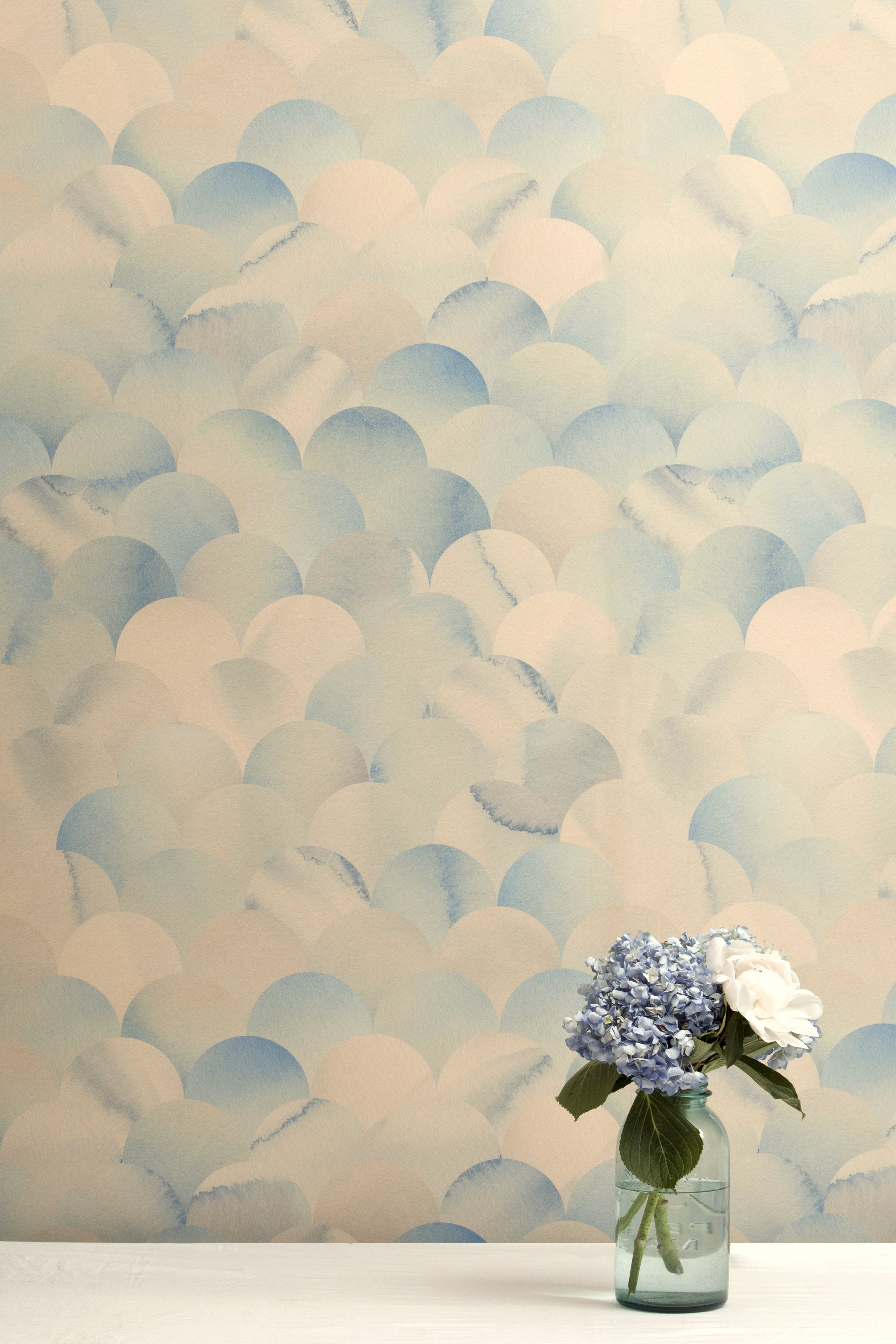 A vase of flowers stands in front of a wall papered in a repeating scalloped print in shades of blue and tan.