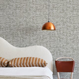 A modernist bed, hanging lamp and chair stand in front of a wall papered in a textural checked print in gray and white.