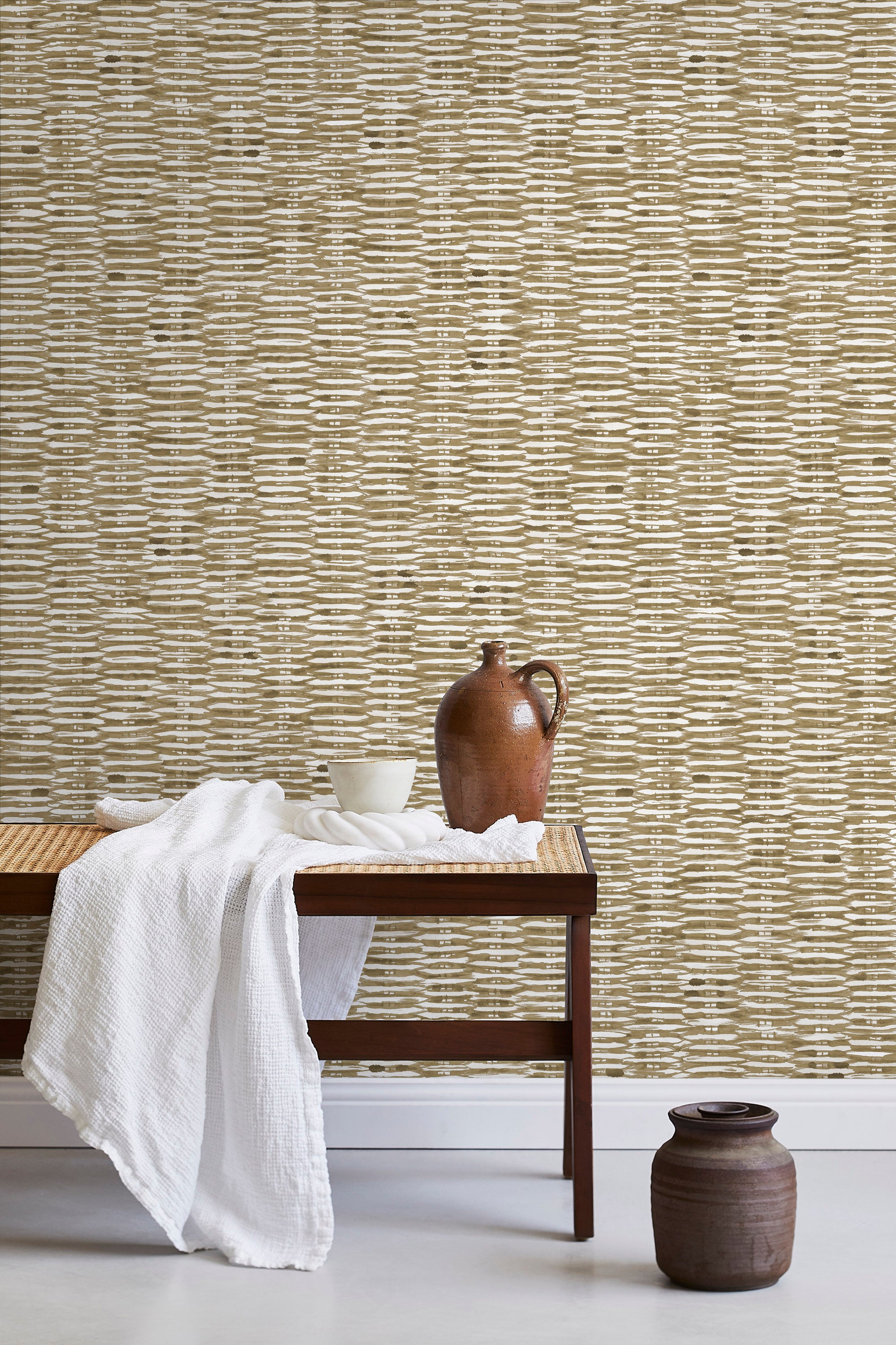 A bench with a towel and clay vases stands in front of a wall papered in a textural checked print in brown and white.