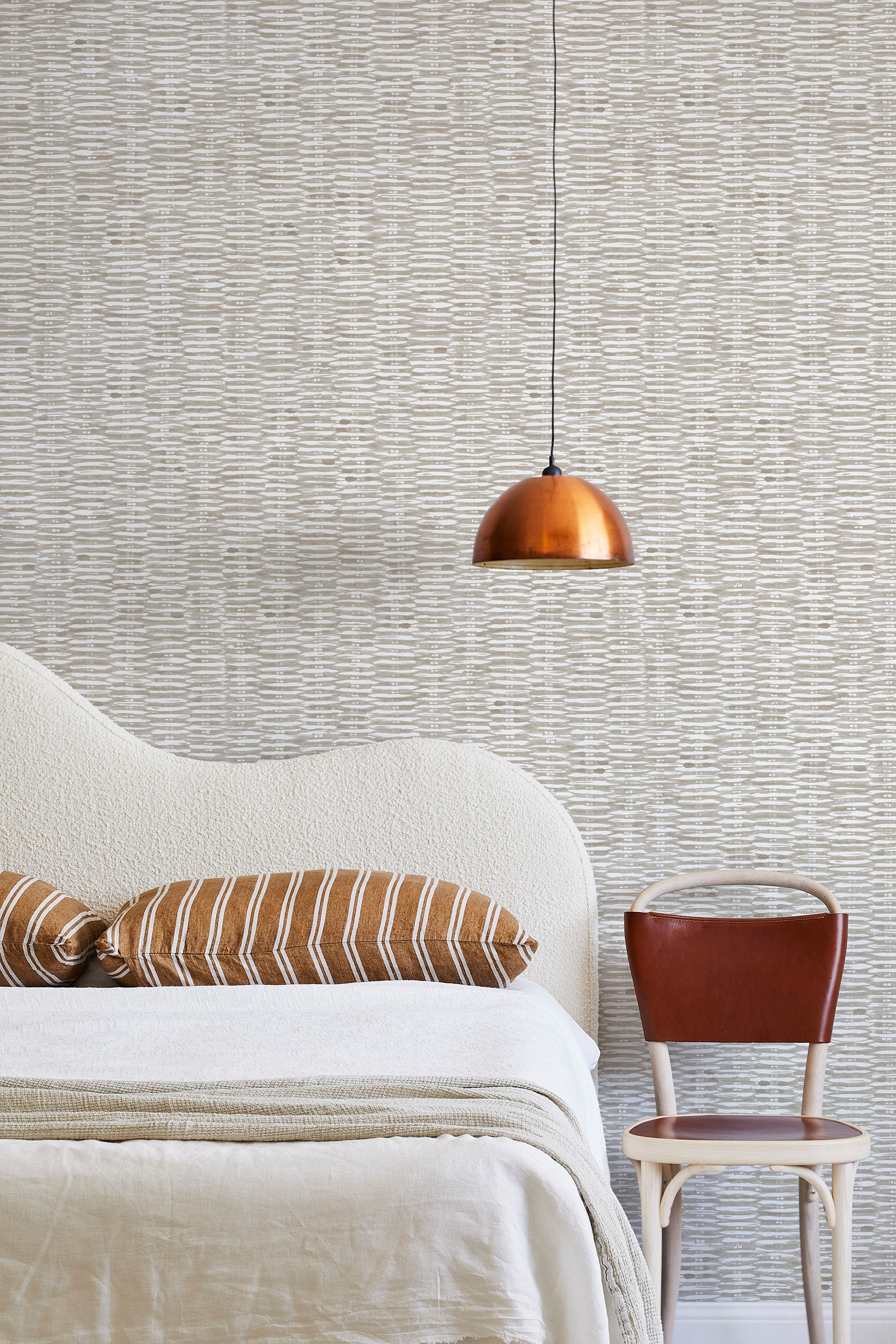A modernist bed, hanging lamp and chair stand in front of a wall papered in a textural checked print in tan and white.
