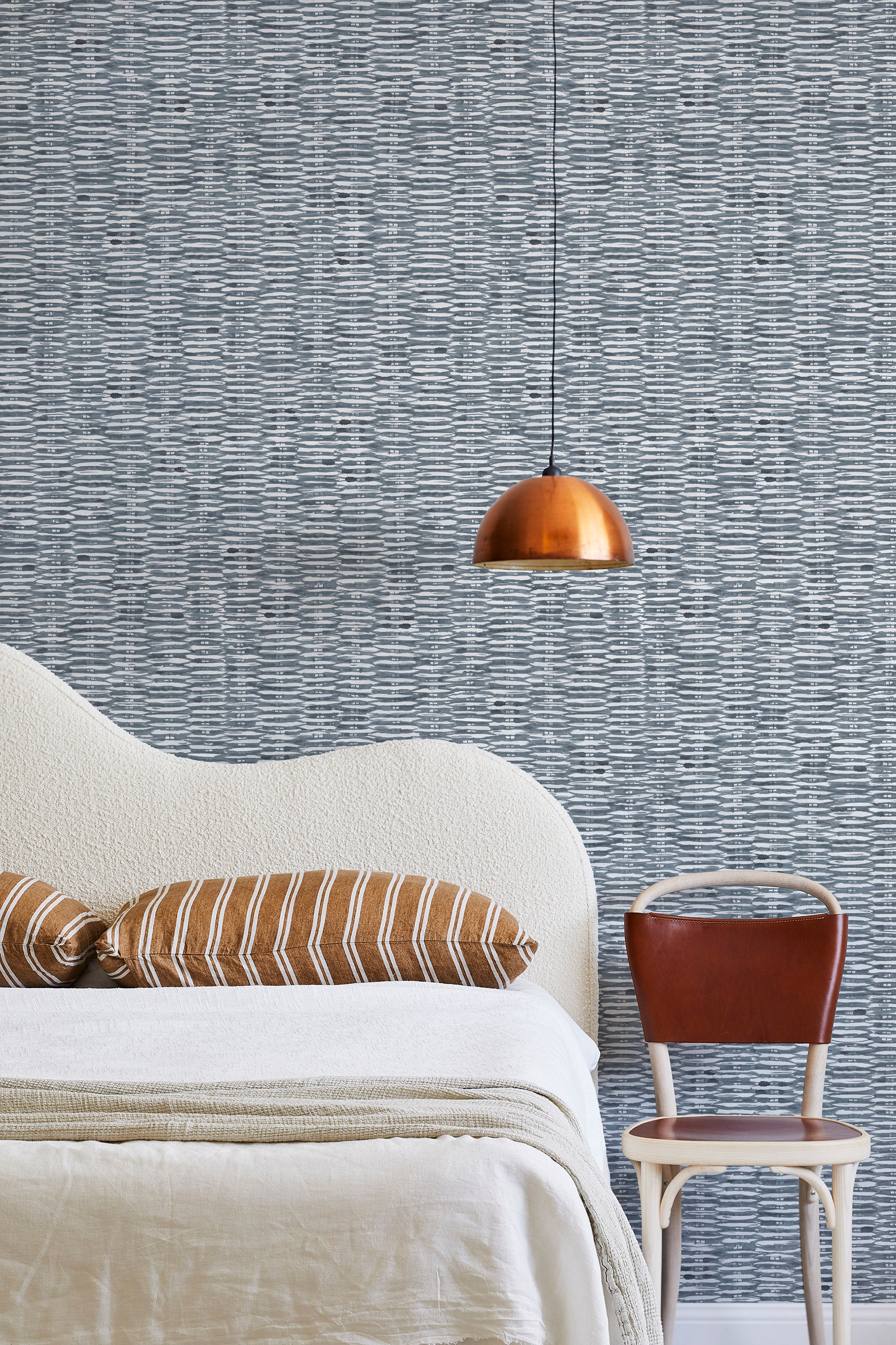 A modernist bed, hanging lamp and chair stand in front of a wall papered in a textural checked print in navy and white.
