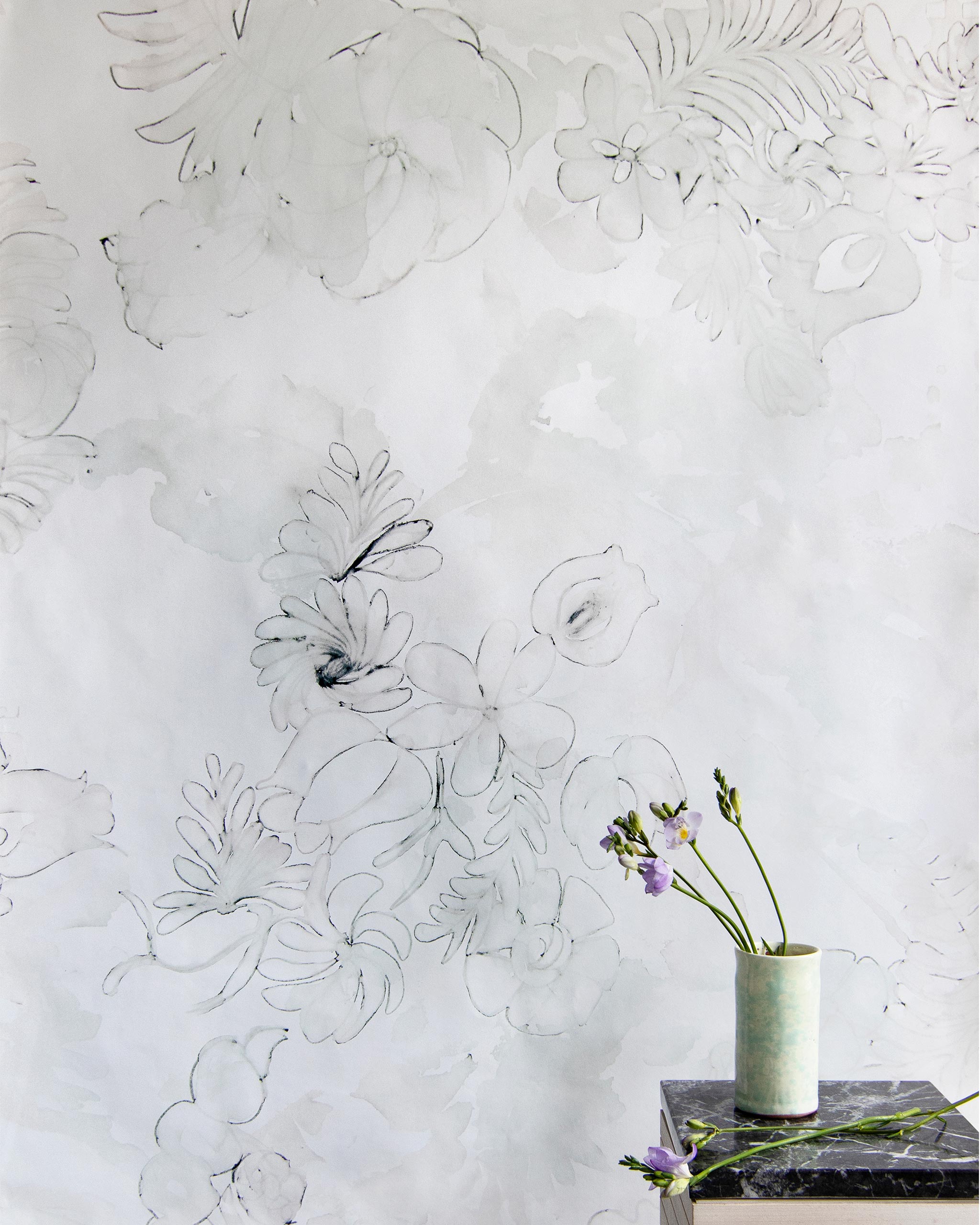 A vase of flowers stands in front of a wall papered in a painterly floral print in pink, charcoal and white.