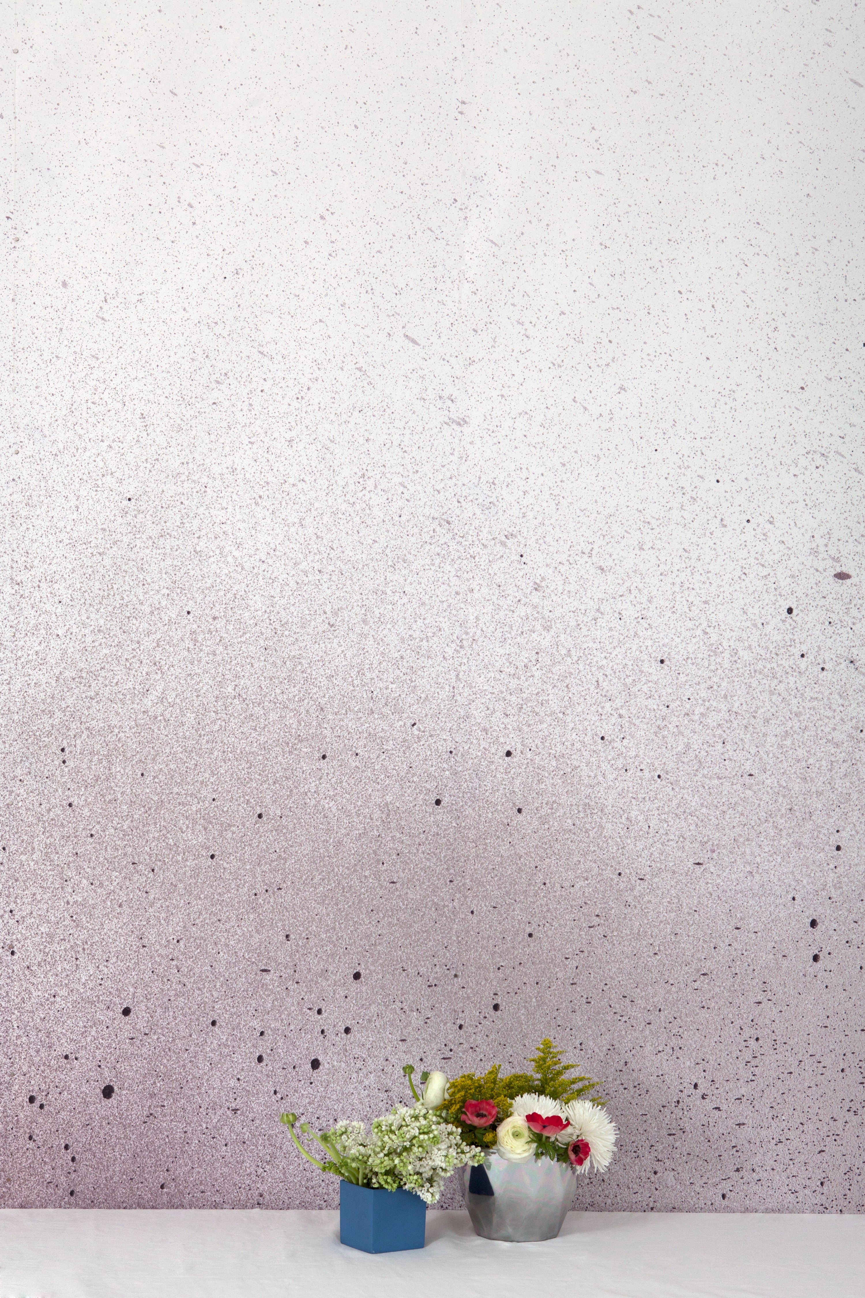 Two vases of flowers stand in front of a wall covered in an abstract ombré paint splatter print in purple, gray and white.