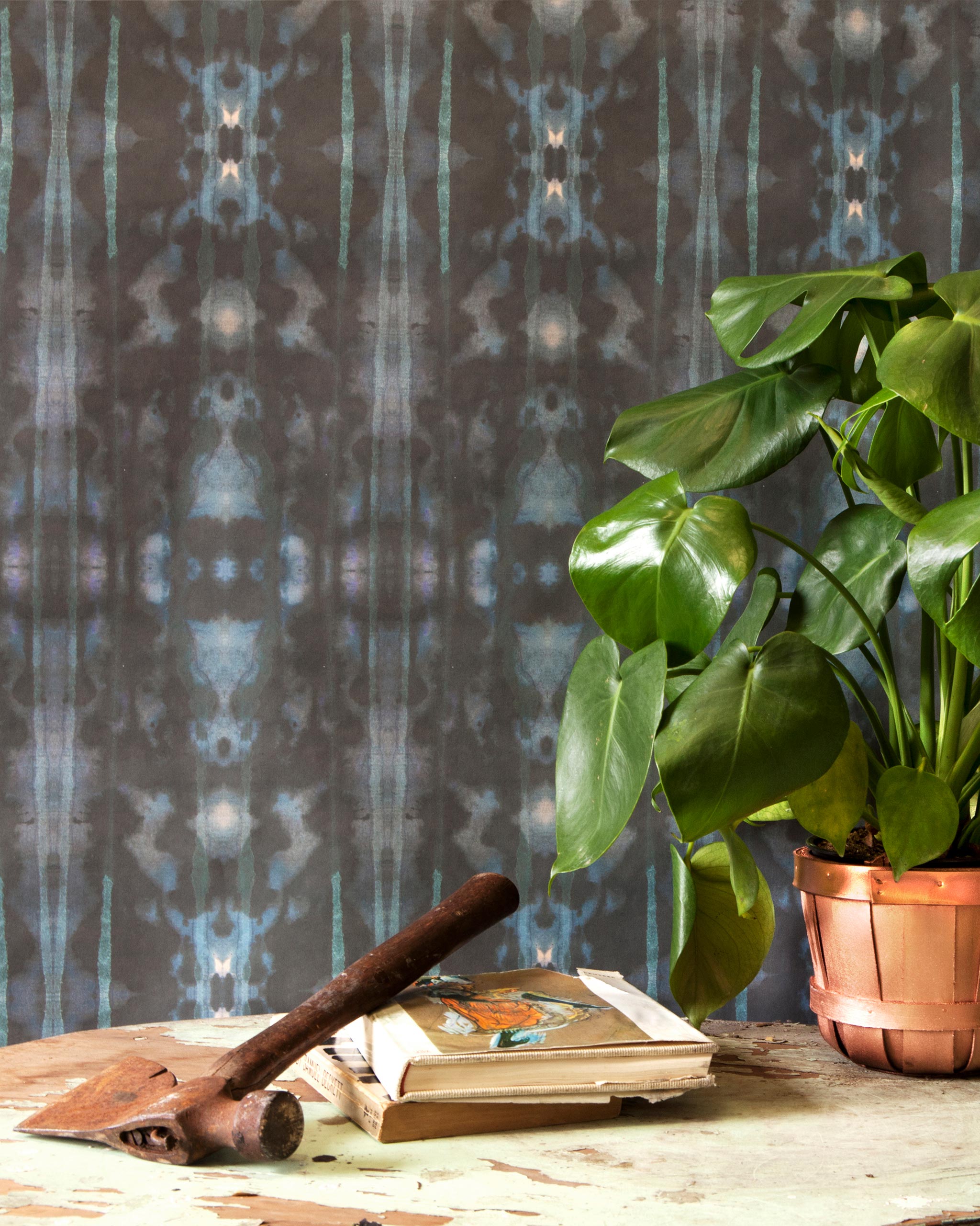 A house plant, some books and a hammer stand in front of a wall papered in a painterly ikat print in turquoise and brown.