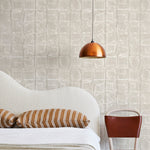 A modernist bed, hanging lamp and chair stand in front of a wall papered in a textural block print in mottled tan.