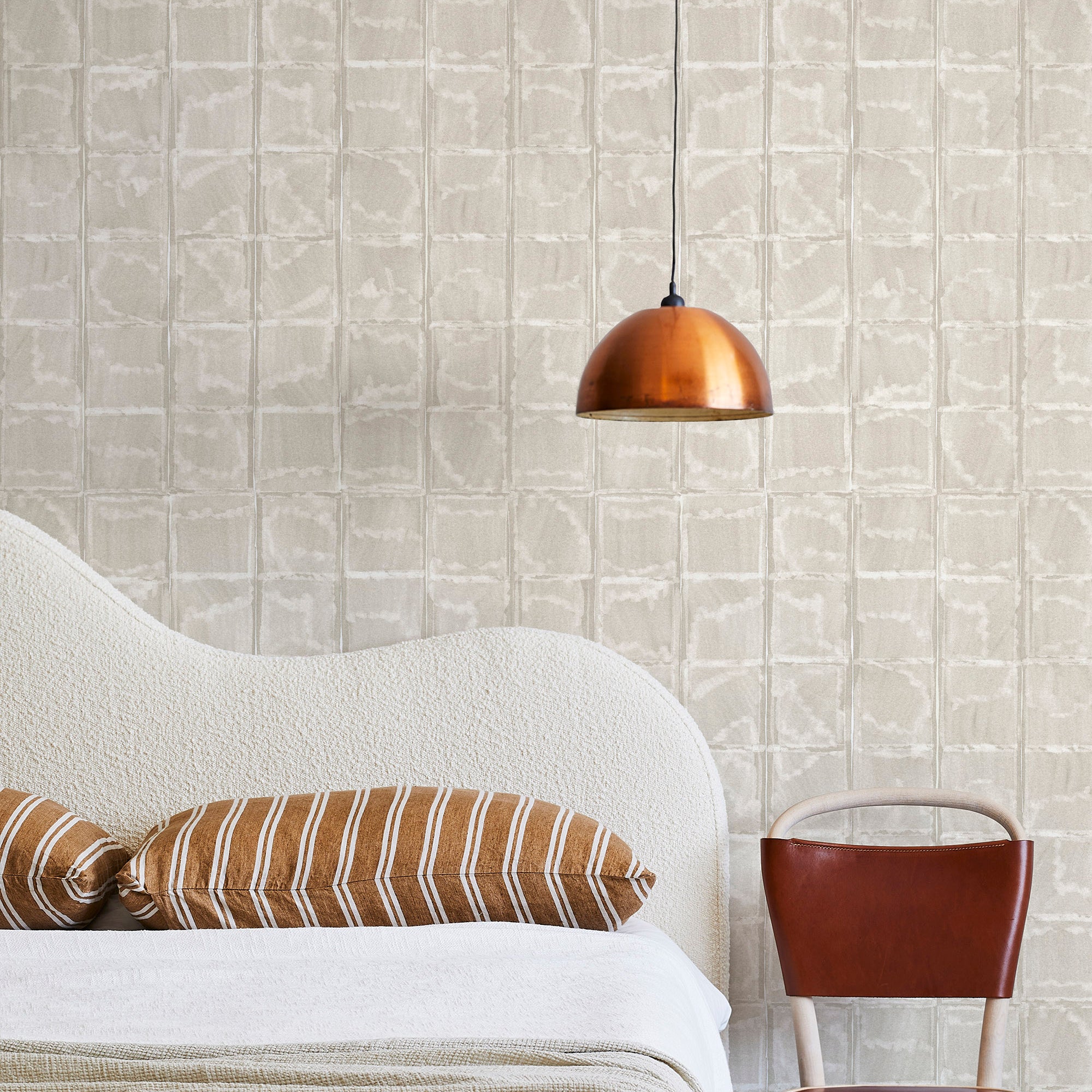 A modernist bed, hanging lamp and chair stand in front of a wall papered in a textural block print in mottled tan.