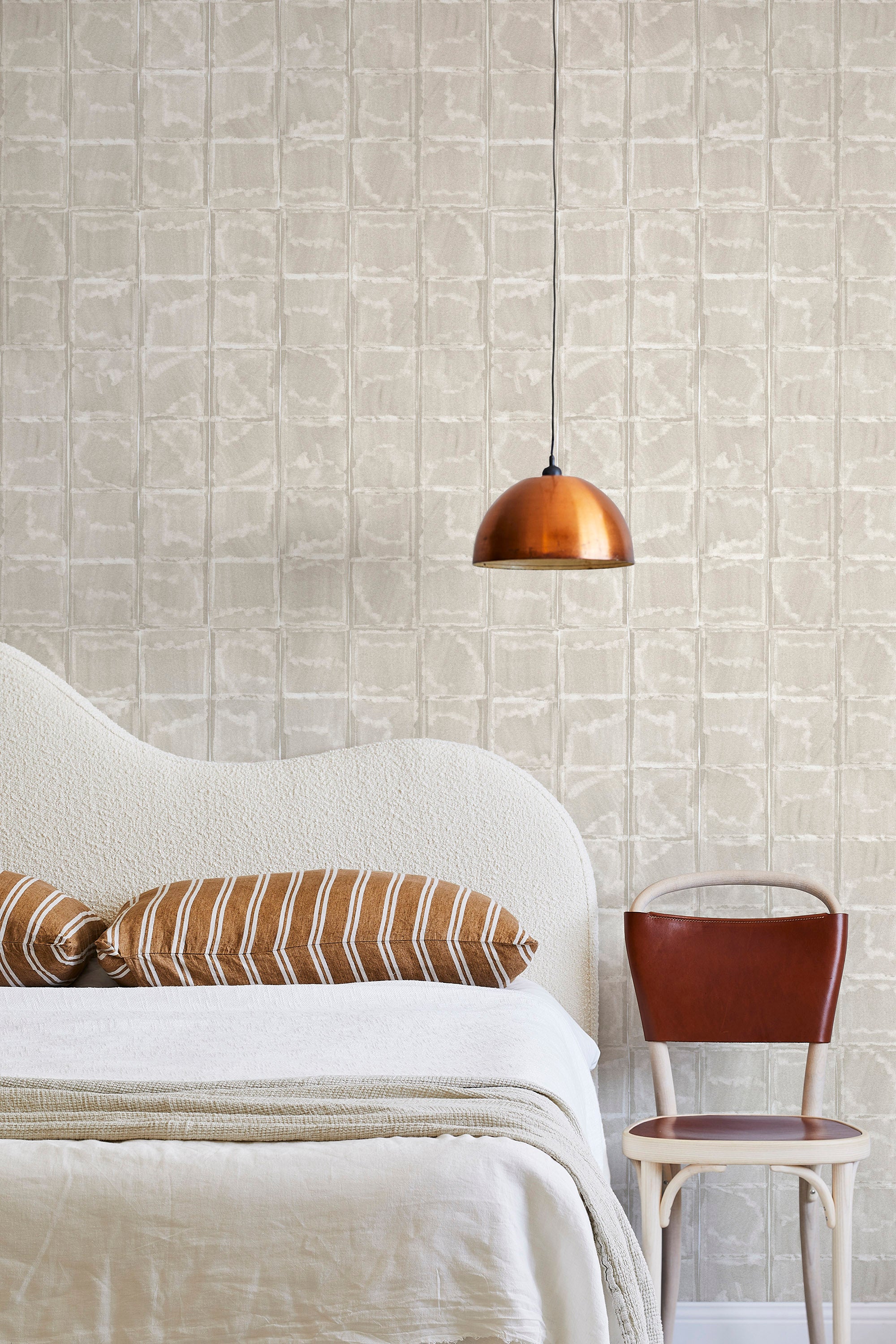 A modernist bed, hanging lamp and chair stand in front of a wall papered in a textural block print in mottled tan.