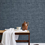 A bench with a towel and clay vases stands in front of a wall papered in a textural block print in mottled blue-black.