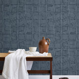 A bench with a towel and clay vases stands in front of a wall papered in a textural block print in mottled blue-black.