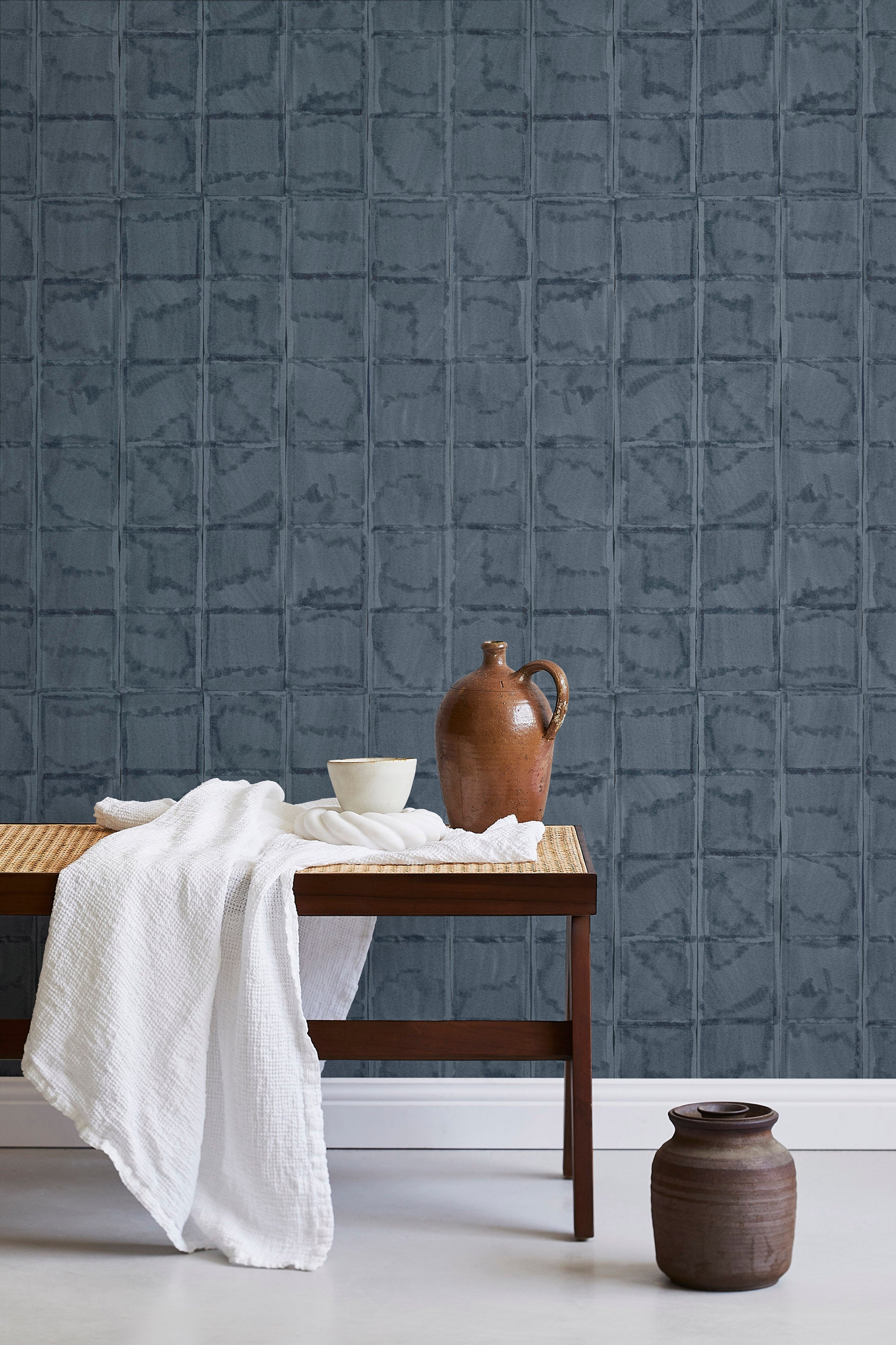 A bench with a towel and clay vases stands in front of a wall papered in a textural block print in mottled blue-black.