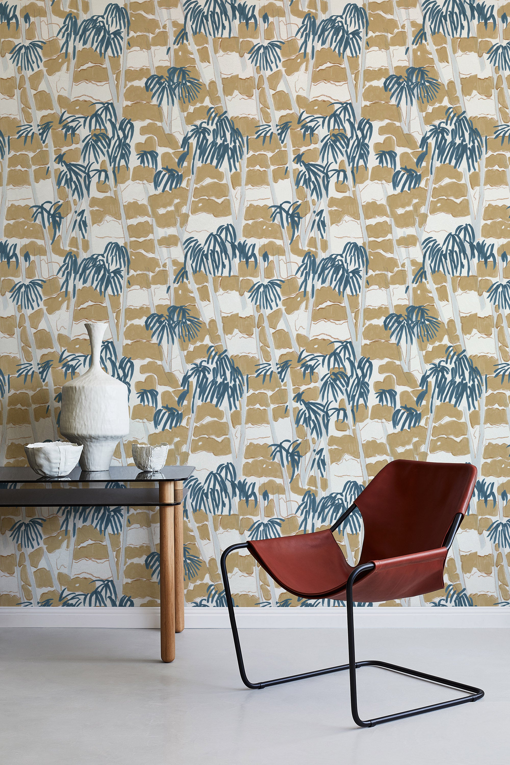 A modernist armchair stands in front of a wall papered in a painterly palm tree print in shades of gray, brown and navy.