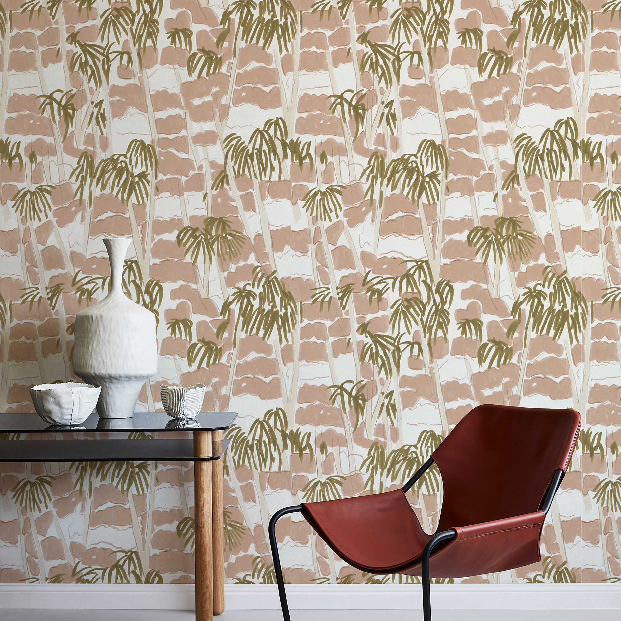 A modernist armchair stands in front of a wall papered in a painterly palm tree print in shades of pink, beige and olive.