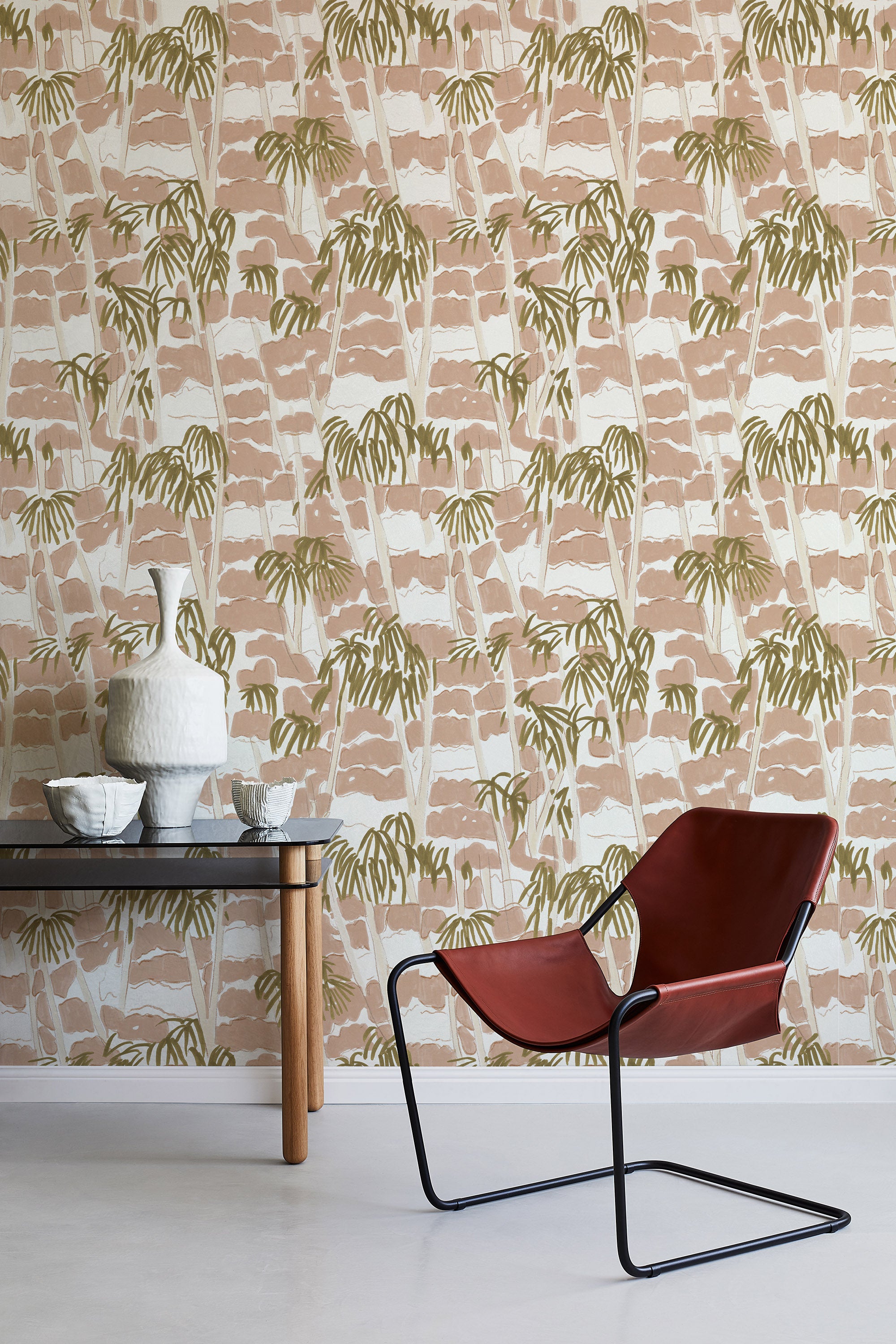 A modernist armchair stands in front of a wall papered in a painterly palm tree print in shades of pink, beige and olive.