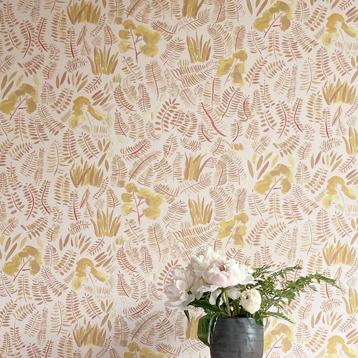 A potted plant stands in front of a wall papered in a playful leaf print in shades of yellow and orange on a white field.