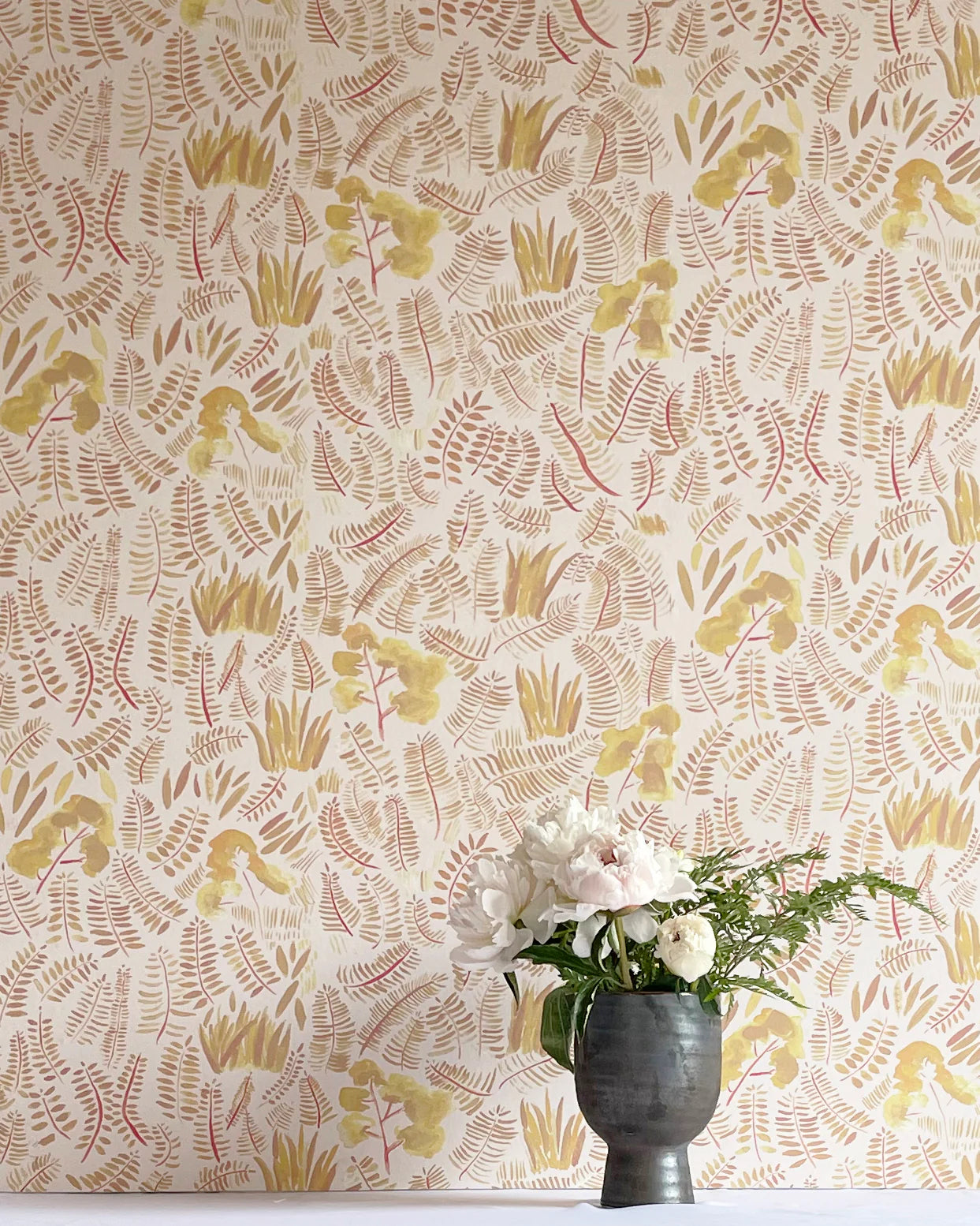 A potted plant stands in front of a wall papered in a playful leaf print in shades of yellow and orange on a white field.