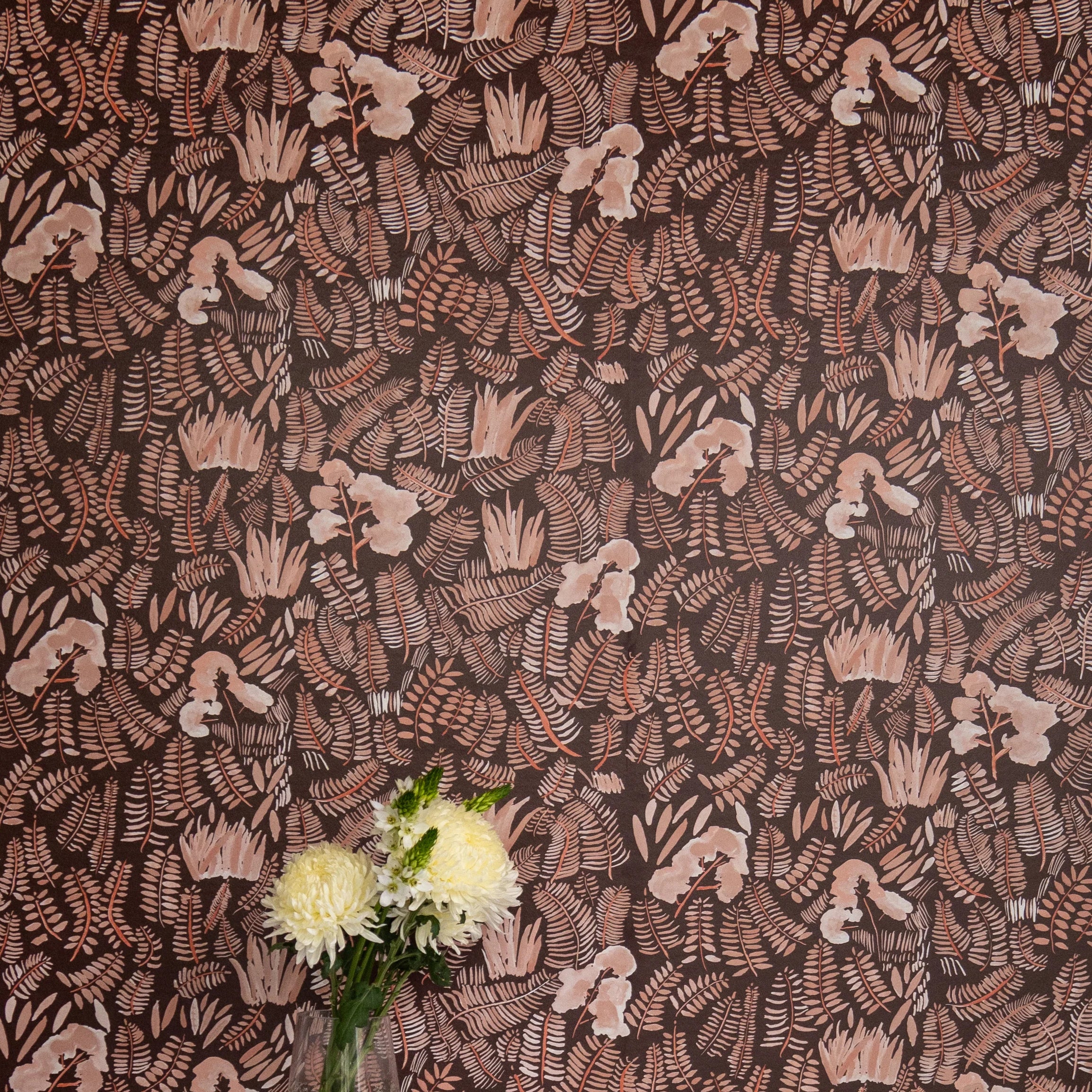 A vase of flowers stands in front of a wall papered in a playful leaf print in shades of orange and coral on a brown field.