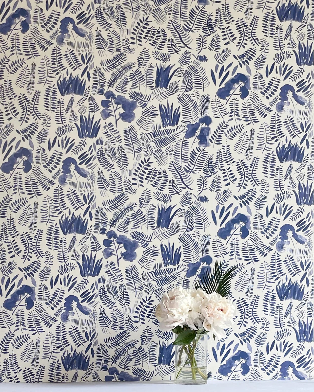 A vase of flowers stands in front of a wall papered in a playful leaf print in shades of blue on a cream field.