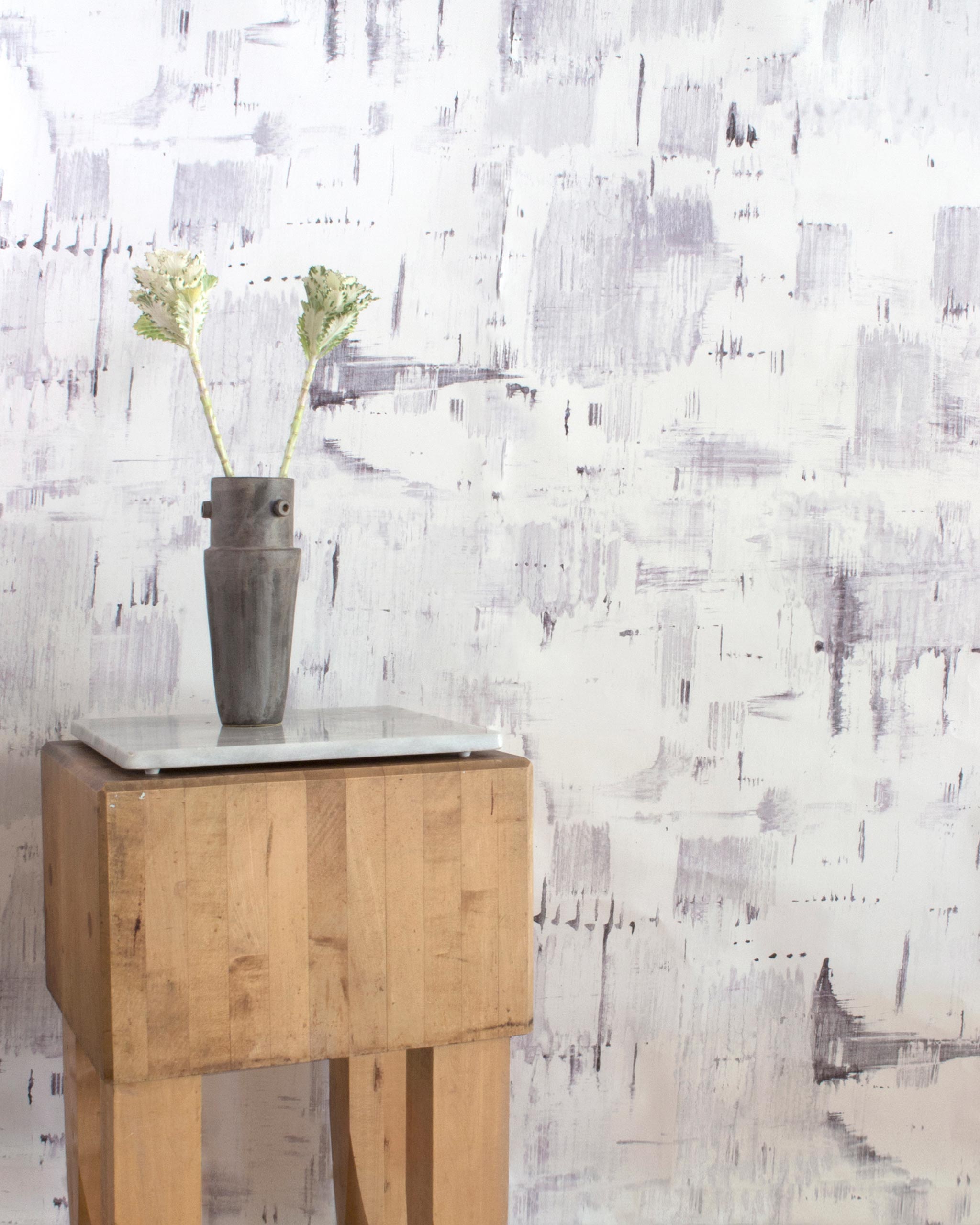 An end table with flowers stands in front of a wall papered in an abstract textural print in shades of gray on a white field.