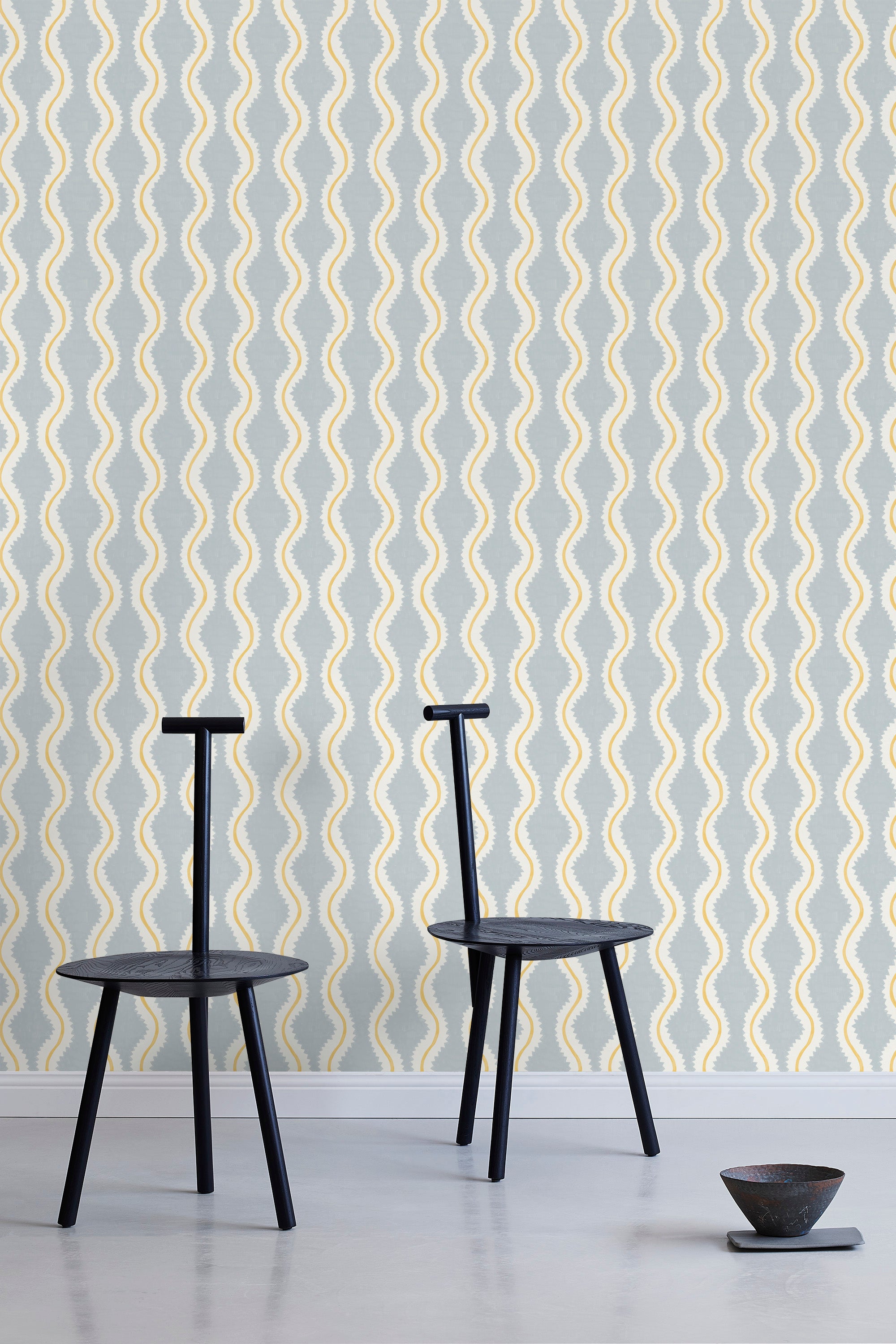 Two minimalist black chairs stand in front of a wall papered in a playful curvy stripe print in white, yellow and light blue.