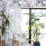 A living space with plants, a window and an accent wall papered in a painterly palm tree print in gray, blue, green and white.