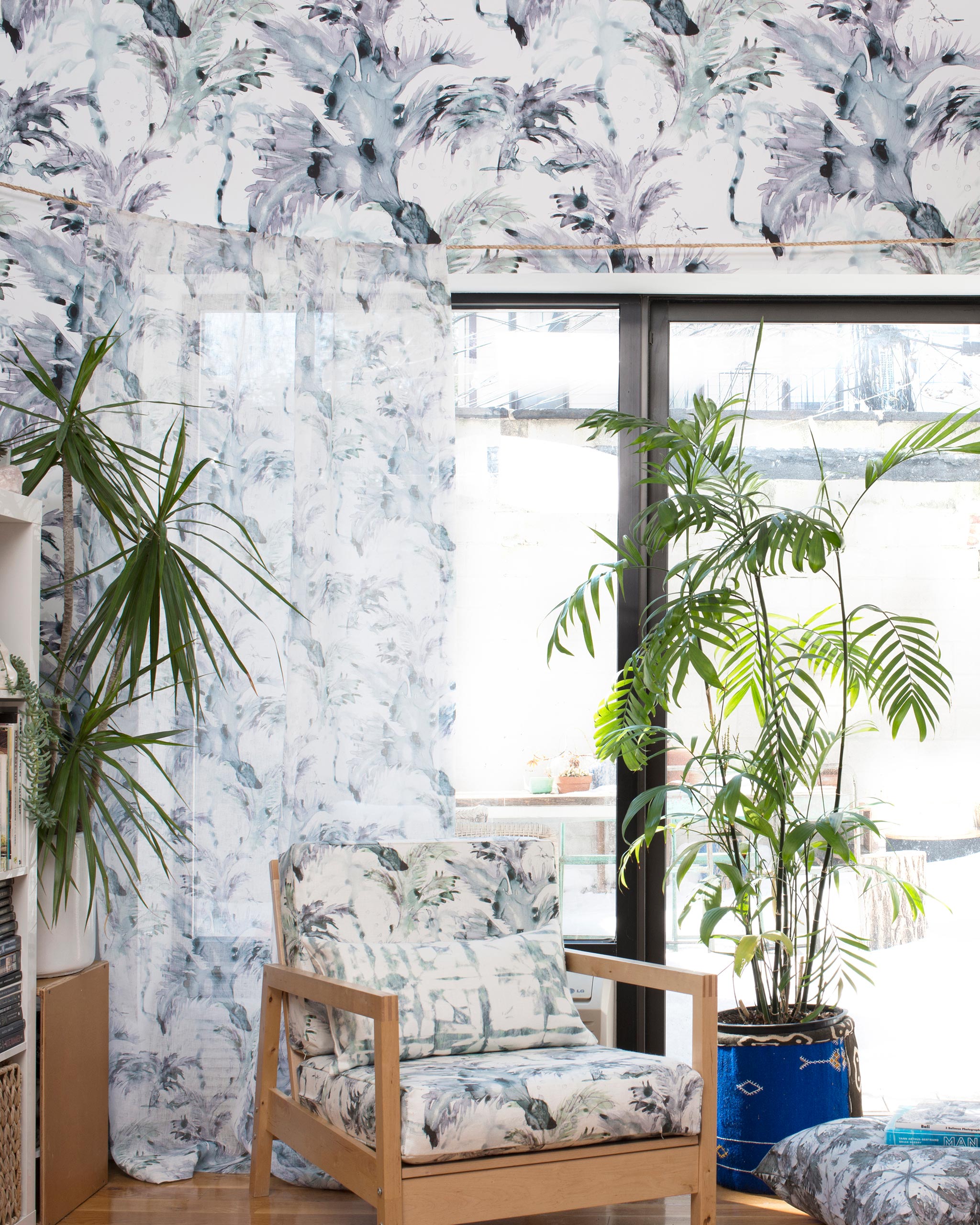 A living space with plants, a window and an accent wall papered in a painterly palm tree print in gray, blue, green and white.