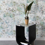 A modernist end table and plant stands in front of a wall papered in an abstract painted print in green, tan and white.