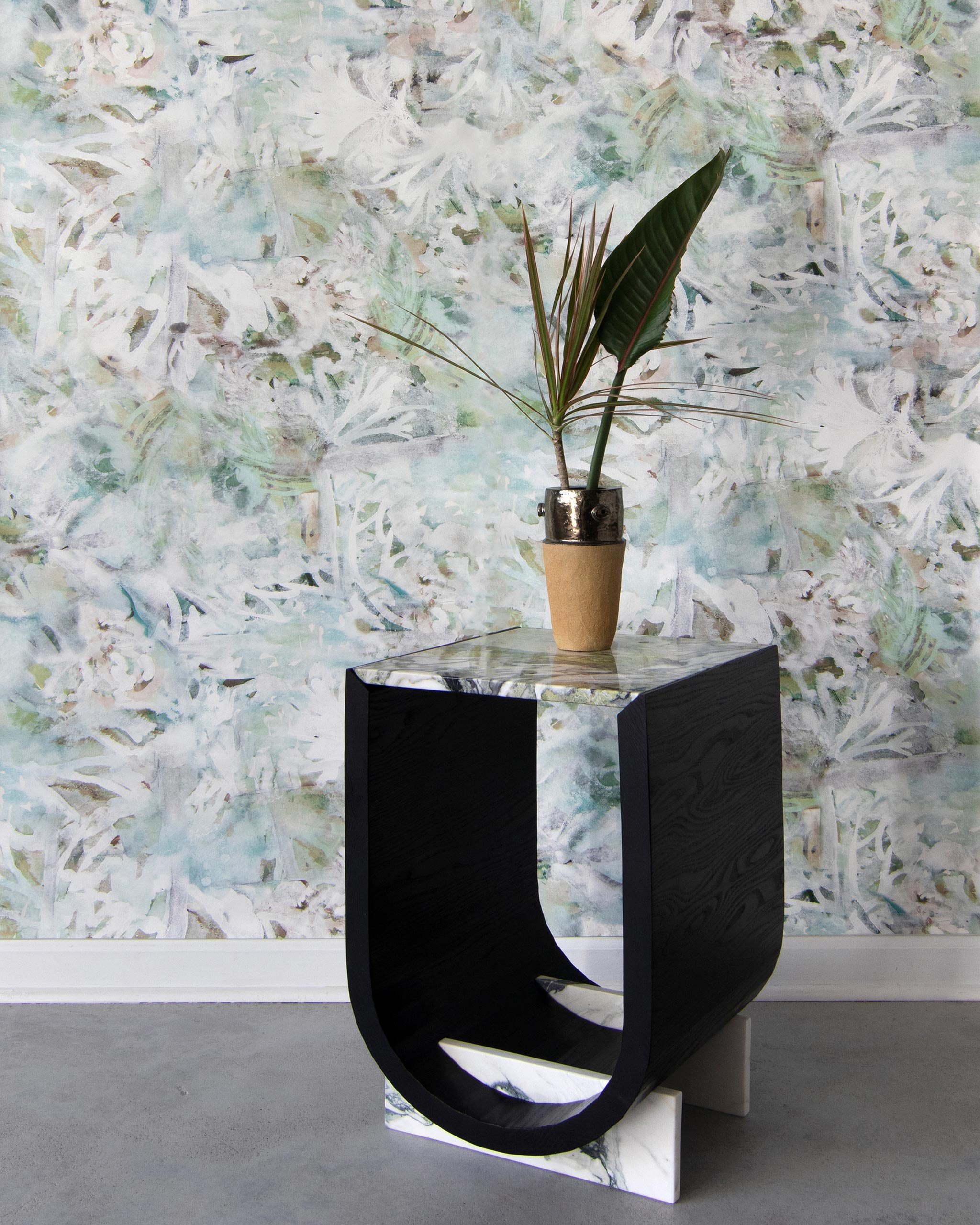 A modernist end table and plant stands in front of a wall papered in an abstract painted print in green, tan and white.