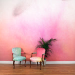 Chairs and a plant stand in front of a wall covered in an abstract ombré paint splatter print in pink, orange and white.