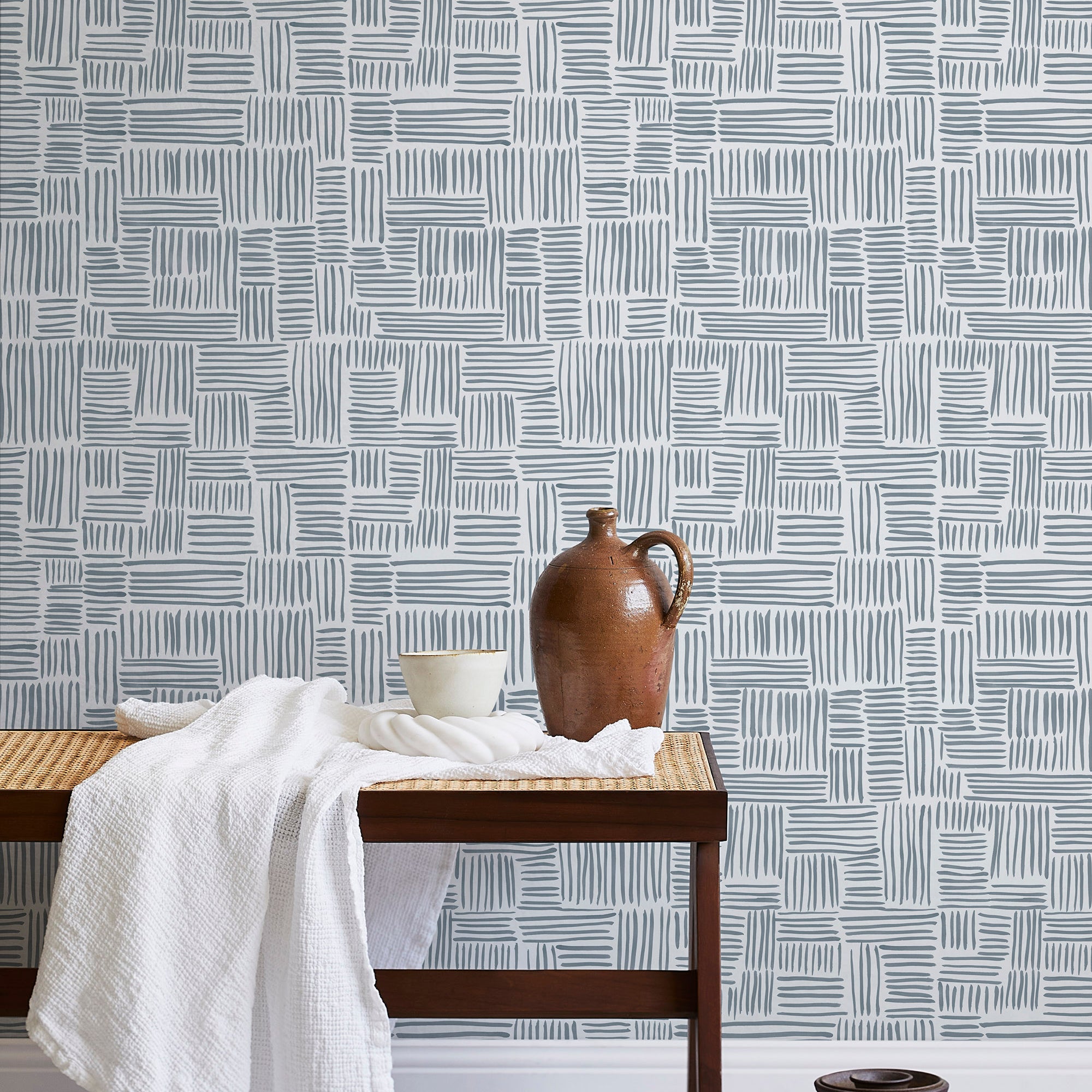 A bench with a towel and clay vases stands in front of a wall papered in a directional dash pattern in blue-gray and white.