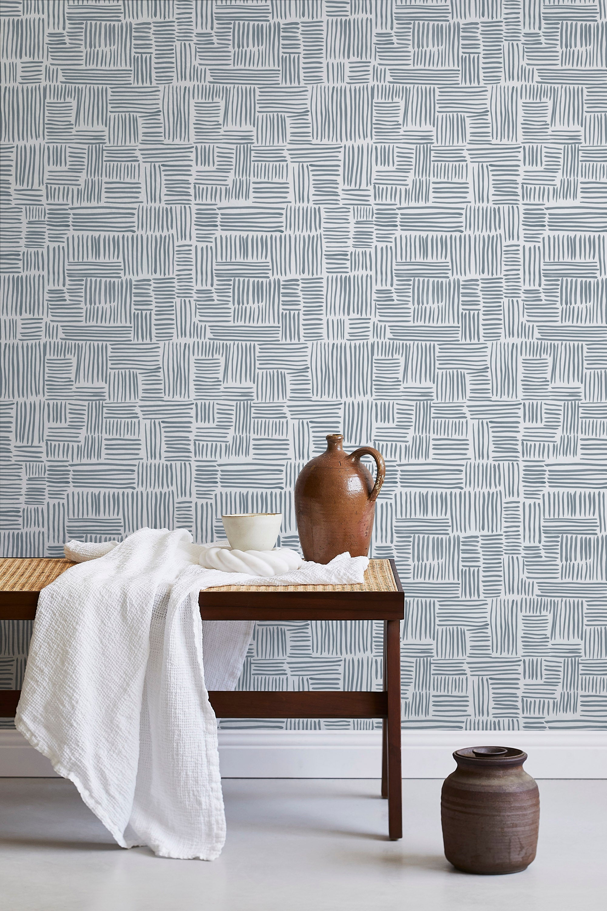 A bench with a towel and clay vases stands in front of a wall papered in a directional dash pattern in blue-gray and white.