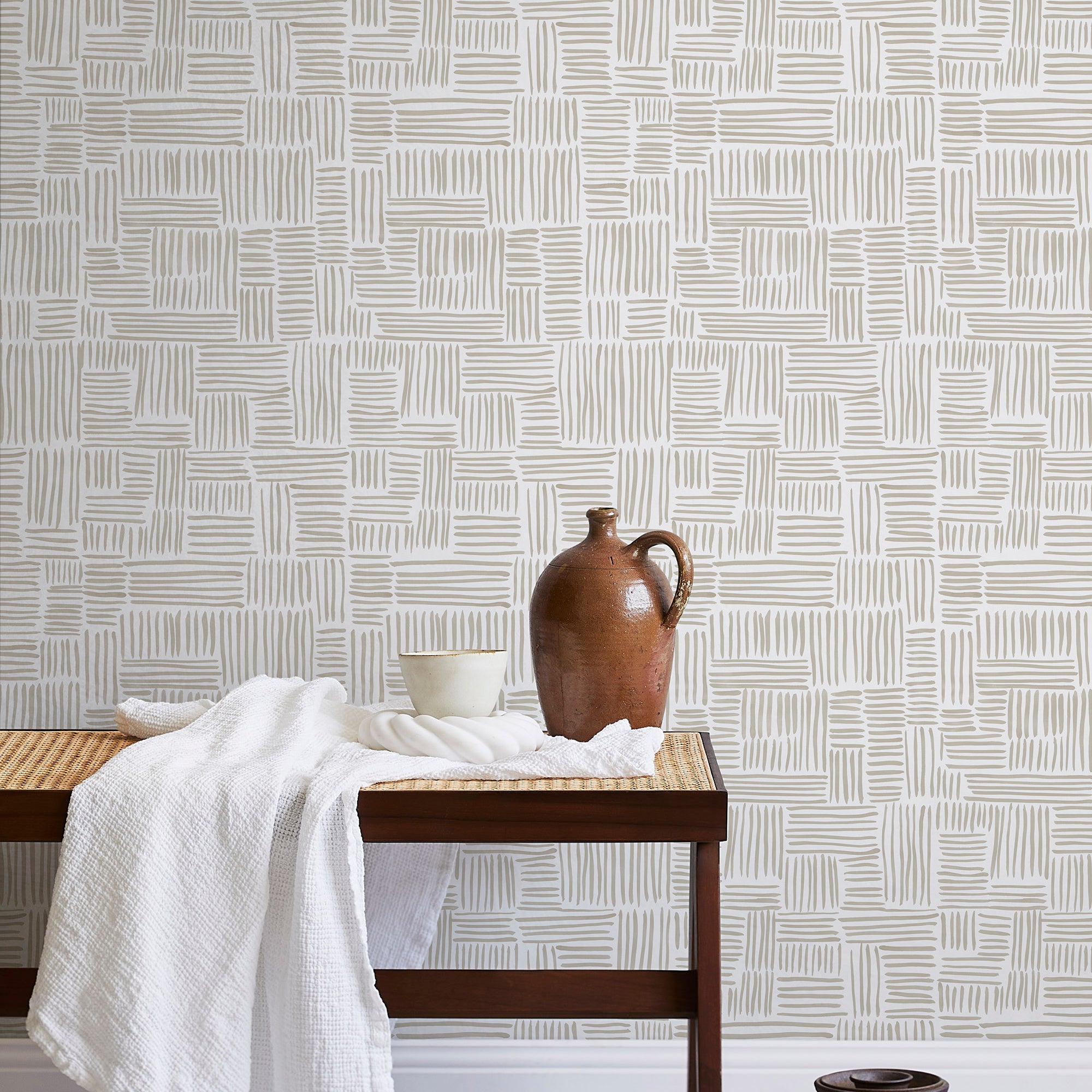 A bench with a towel and clay vases stands in front of a wall papered in a directional dash pattern in cream and white.