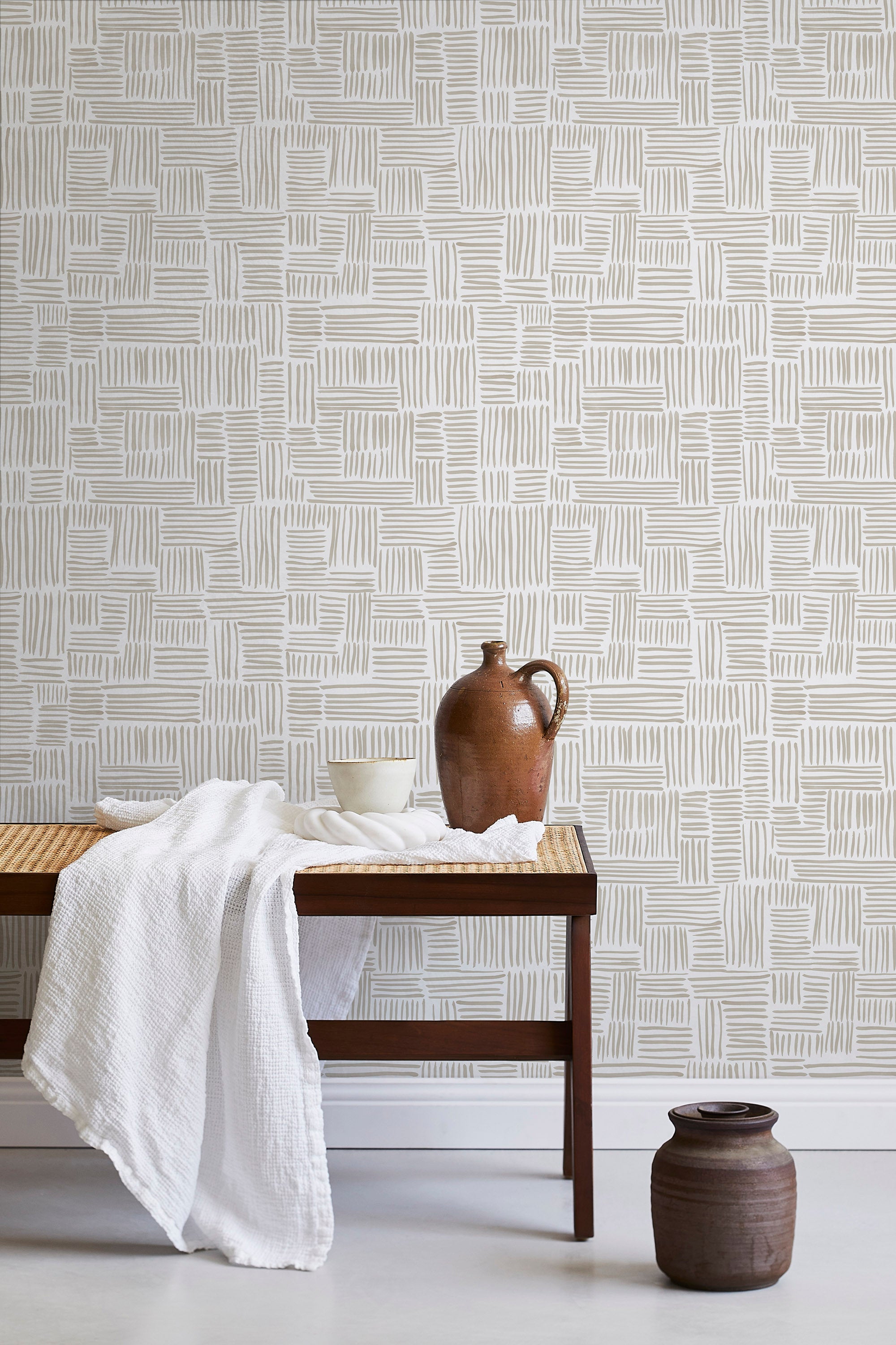 A bench with a towel and clay vases stands in front of a wall papered in a directional dash pattern in cream and white.