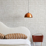 A modernist bed, hanging lamp and chair stand in front of a wall papered in a directional dash pattern in white and cream.