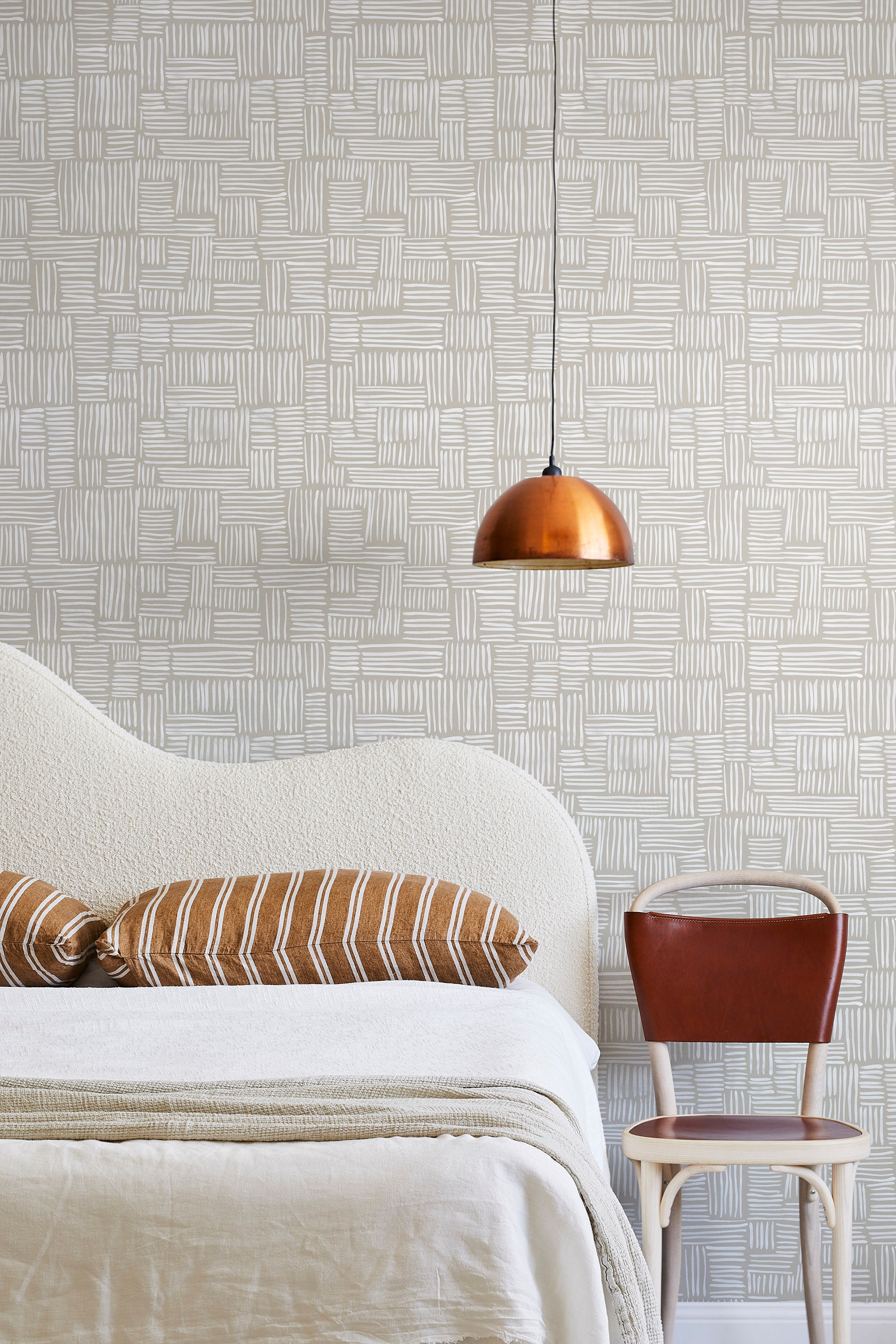 A modernist bed, hanging lamp and chair stand in front of a wall papered in a directional dash pattern in white and cream.