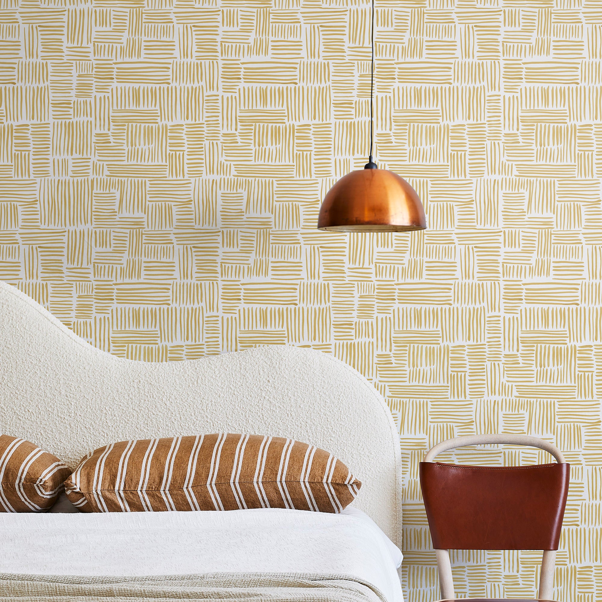 A modernist bed, hanging lamp and chair stand in front of a wall papered in a directional dash pattern in yellow and white.