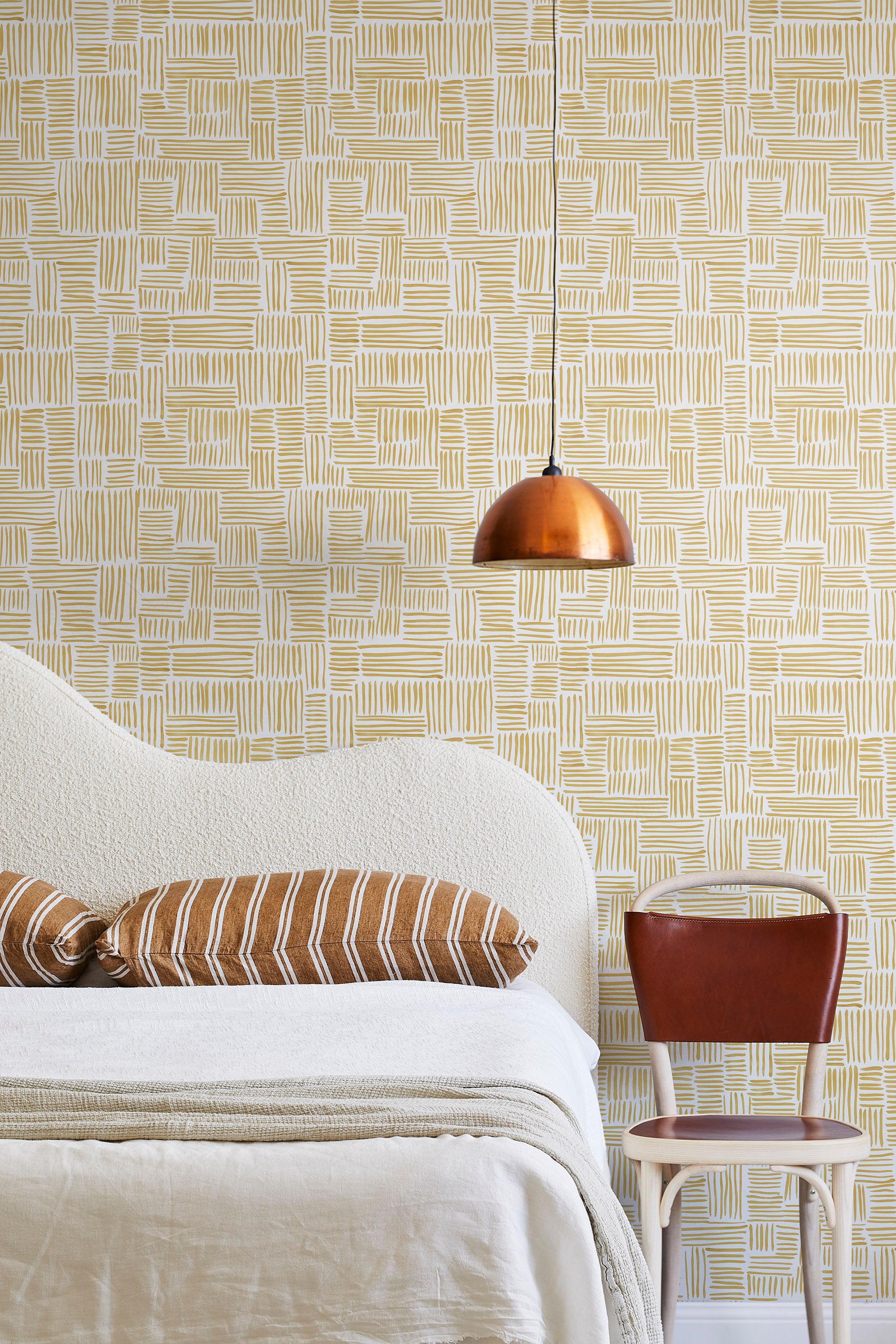 A modernist bed, hanging lamp and chair stand in front of a wall papered in a directional dash pattern in yellow and white.